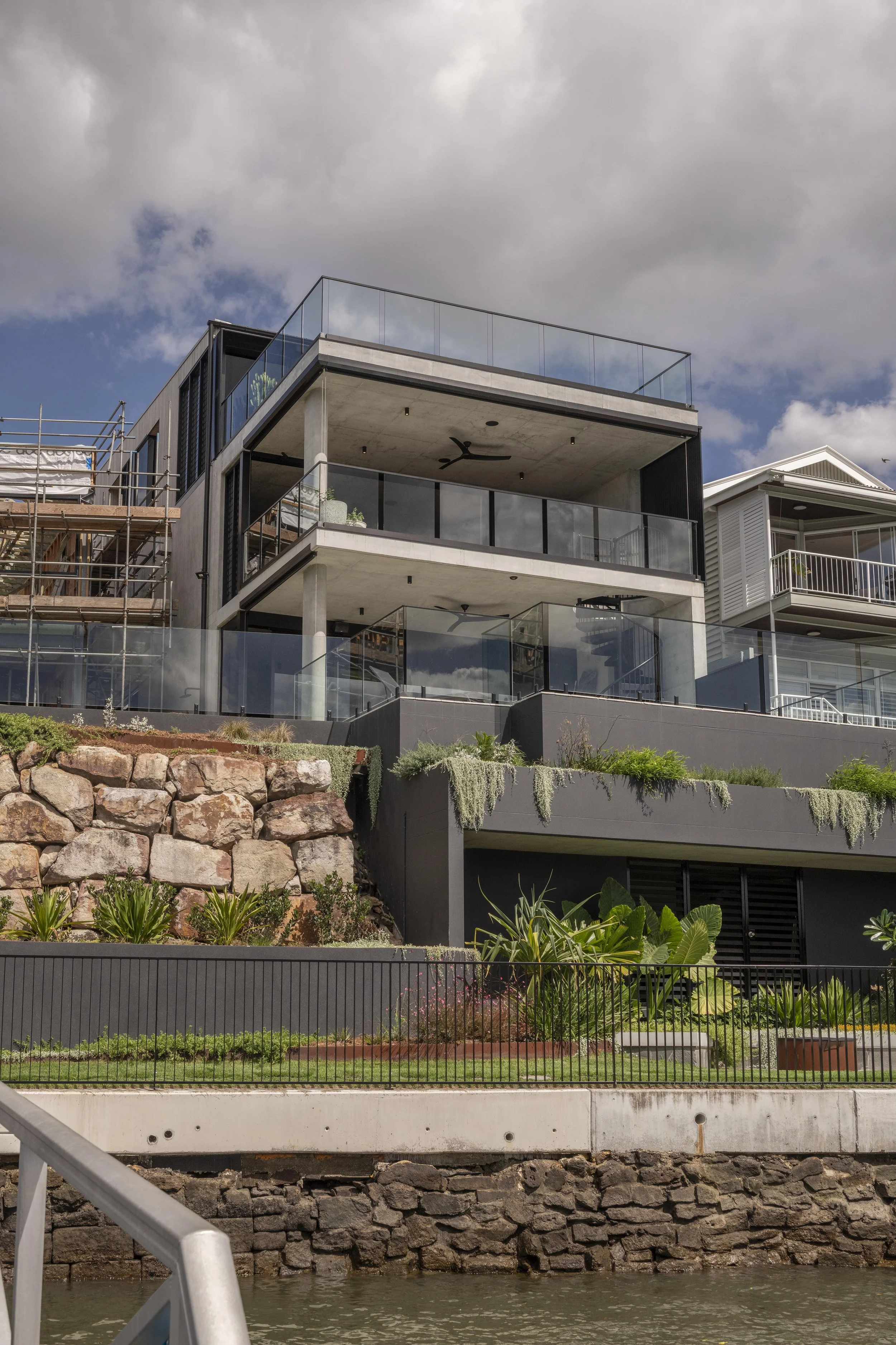 This image showcases a completed knock‑down and rebuild project by Allen Brothers Construction — a multi‑level architectural home positioned on a waterfront site. The new structure features expansive glazing, minimalist concrete forms and layered bal