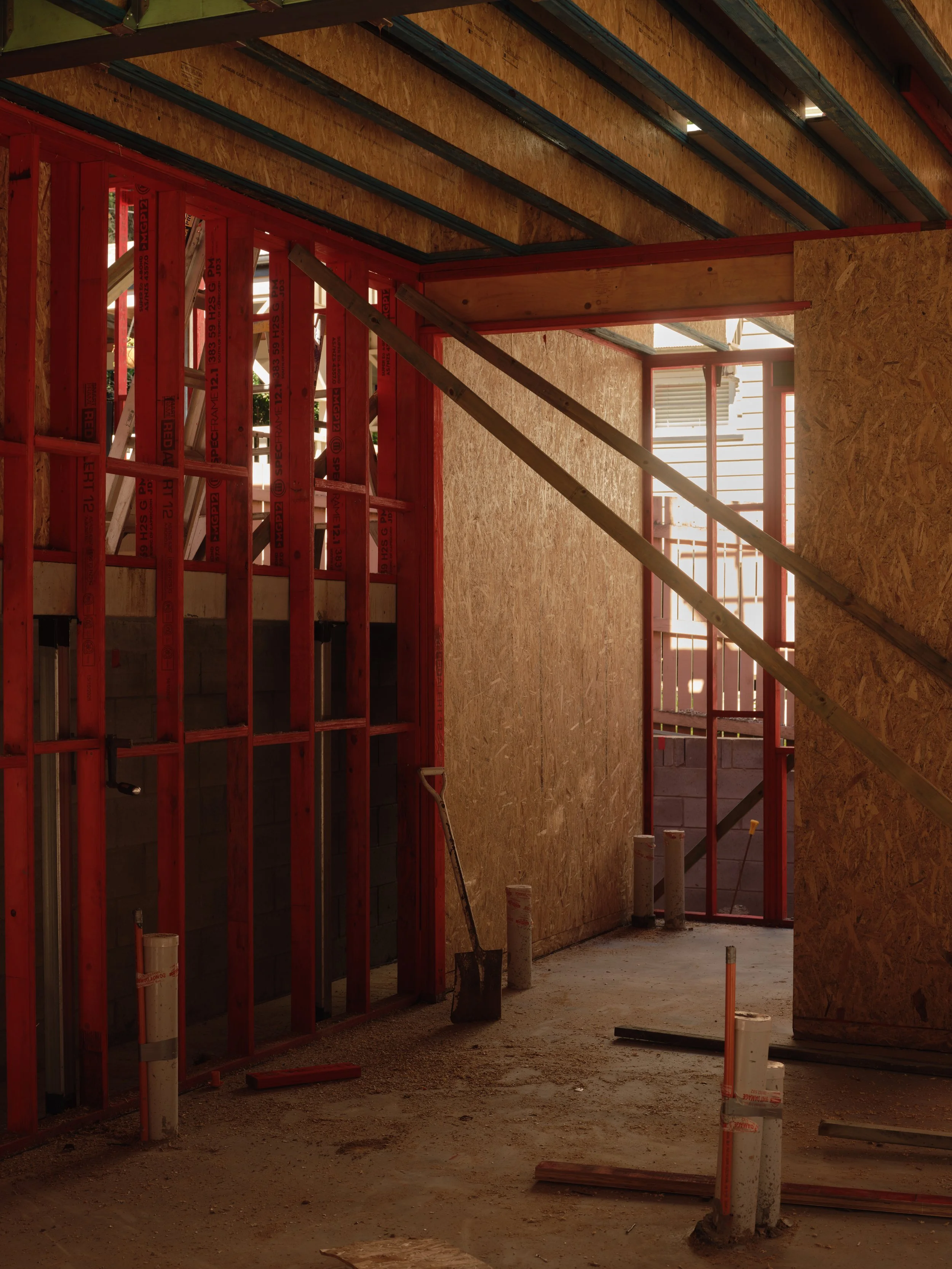 Internal framing stage of Project 135 showing red‑painted timber framing, OSB wall panels, exposed ceiling beams, and plumbing rough‑ins installed through the concrete slab.