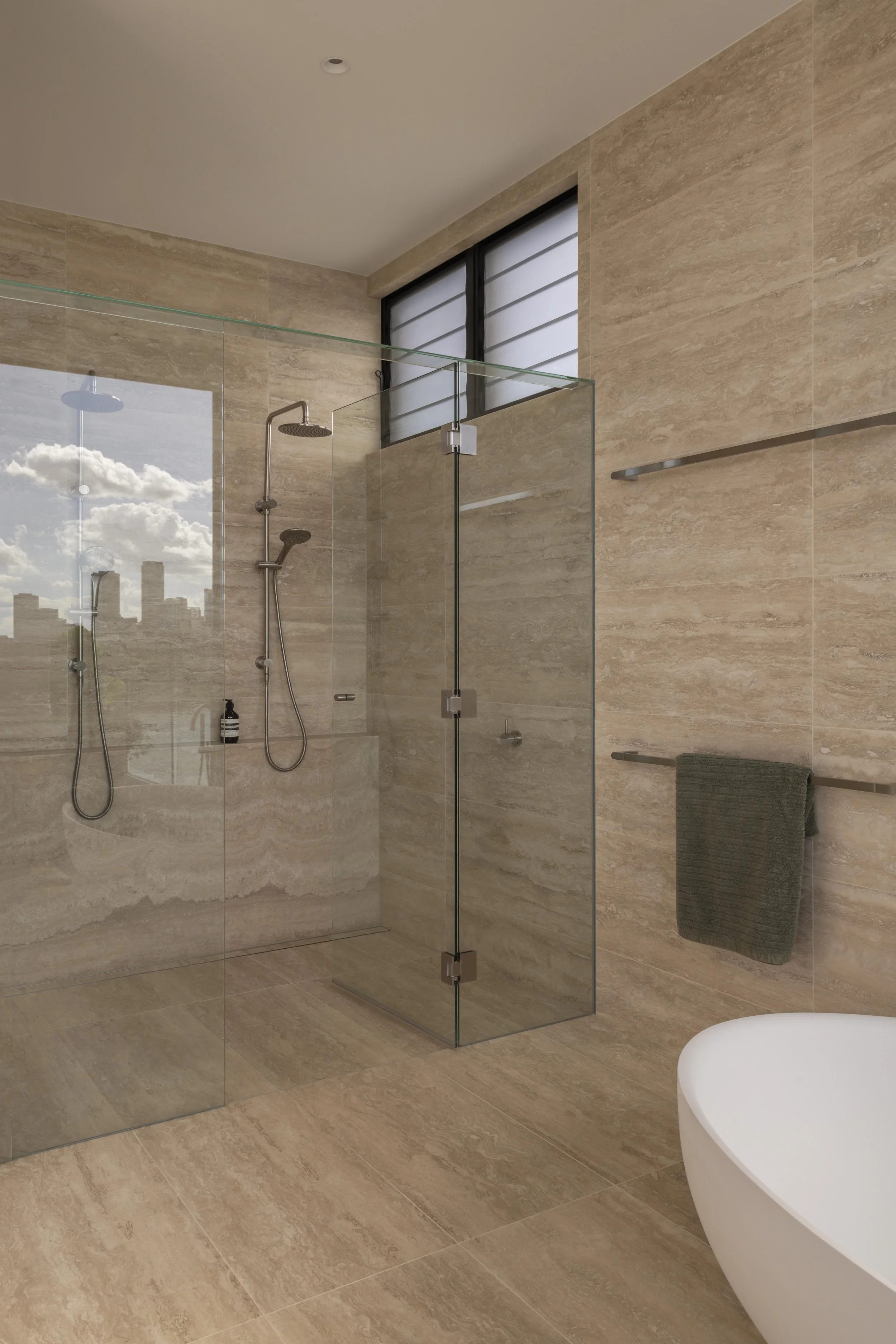 This luxurious bathroom at Project124 features a frameless glass shower with dual showerheads, warm stone surfaces, and soft natural light filtering through horizontal louvred windows. A sculptural freestanding bath sits just beyond the shower zone, 