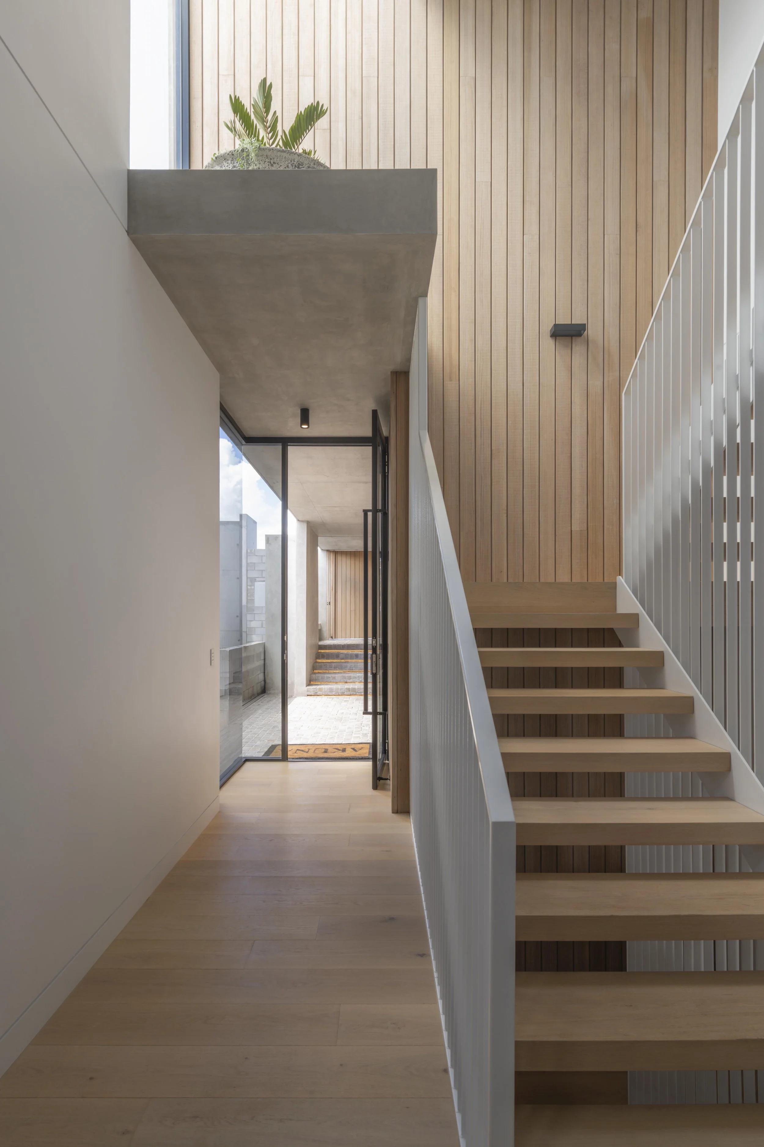 This light‑filled circulation space at Project124 features a minimalist timber staircase with slender white balustrades, complemented by warm vertical timber panelling and crisp white walls. A concrete overhang introduces a touch of raw texture, supp