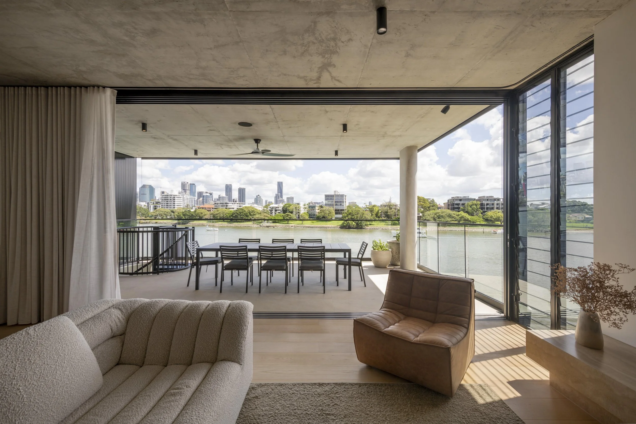 This open living space at Project124 flows seamlessly onto a covered riverside terrace through full‑height sliding glass doors, framing uninterrupted views of the water and city skyline. Soft, textured furnishings, warm timber flooring, and a neutral