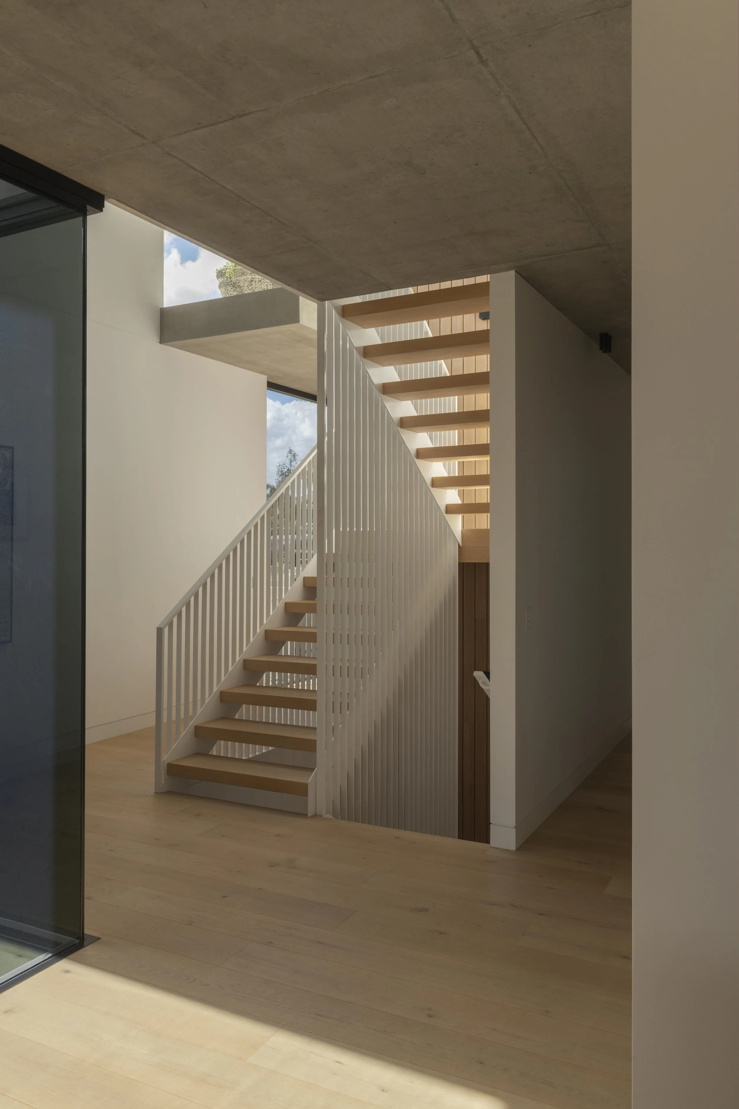 This light‑filled circulation space at Project124 features a minimalist timber staircase with slender white balustrades, soft natural light, and a warm timber floor that continues throughout the home. Exposed concrete overhead adds a subtle industria