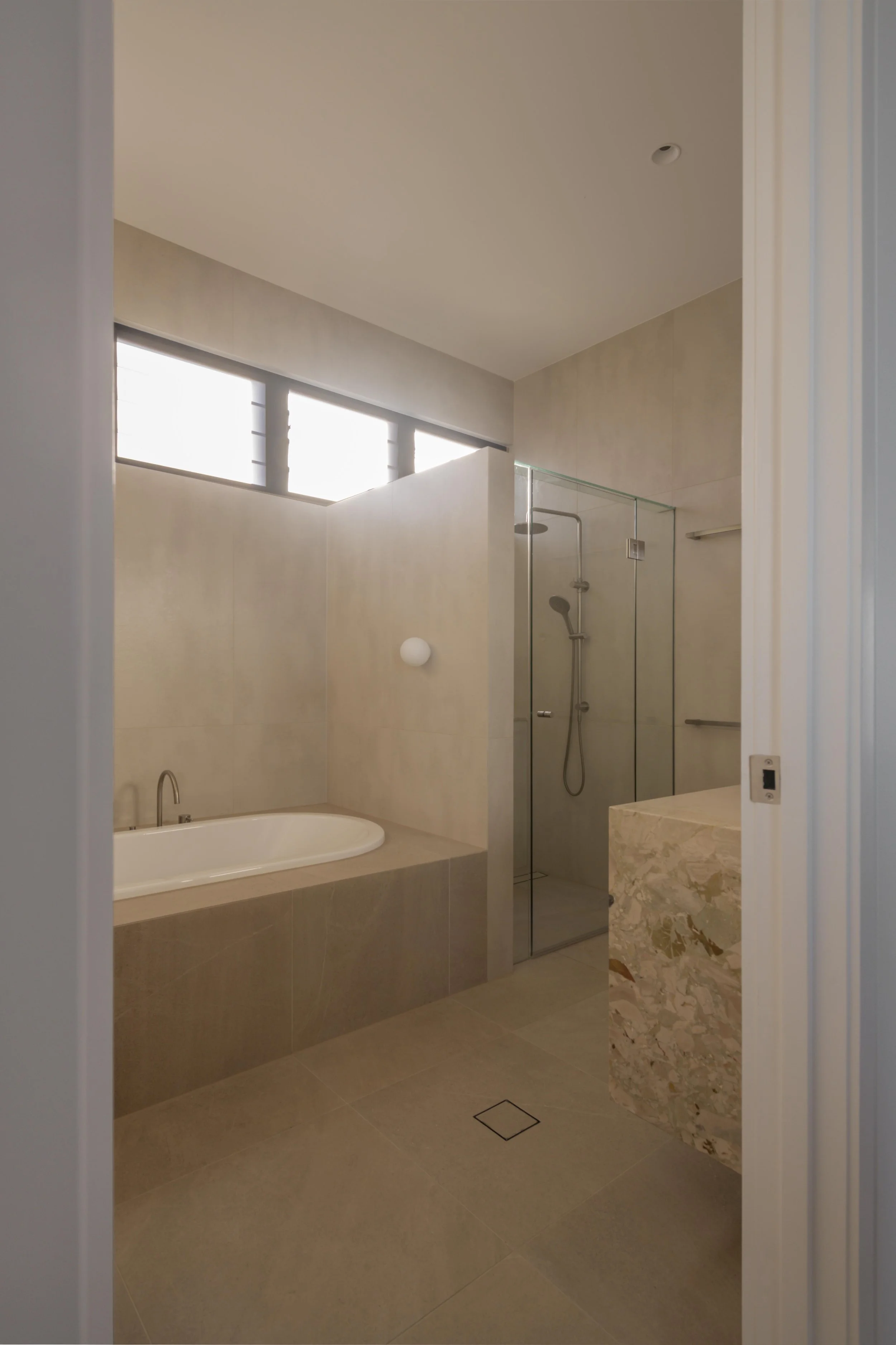 This serene bathroom at Project124 features a sculpted built‑in bath, frameless glass shower, and a warm, neutral palette of large‑format tiles and stone. Soft natural light filters through the louvred windows, enhancing the minimalist detailing and 
