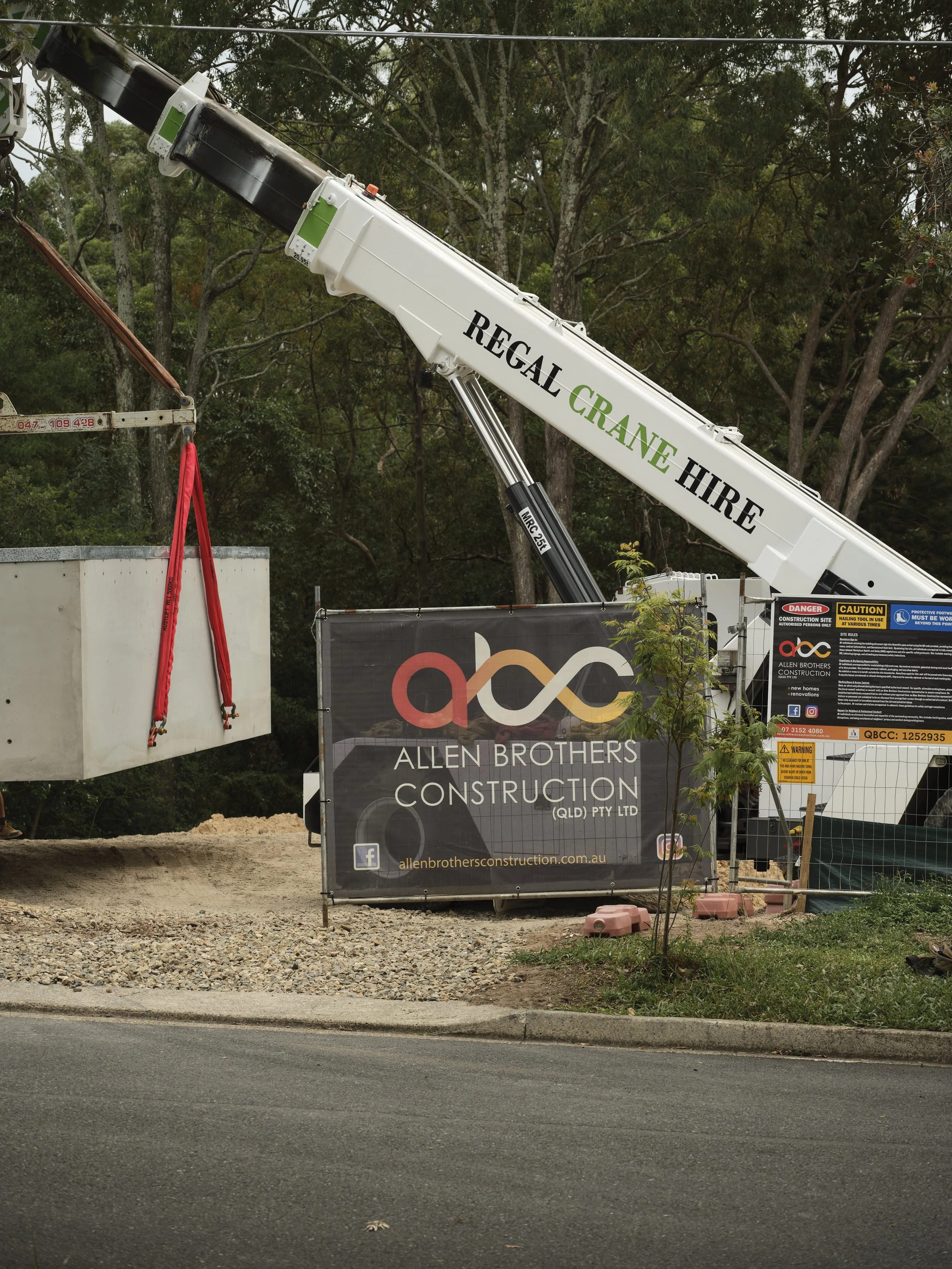 A mobile crane is lifting the precast concrete Plungie pool into position on the construction site. The pool is suspended mid‑air by heavy‑duty lifting chains, guided carefully by the site crew below. The surrounding area shows the early landscaping 