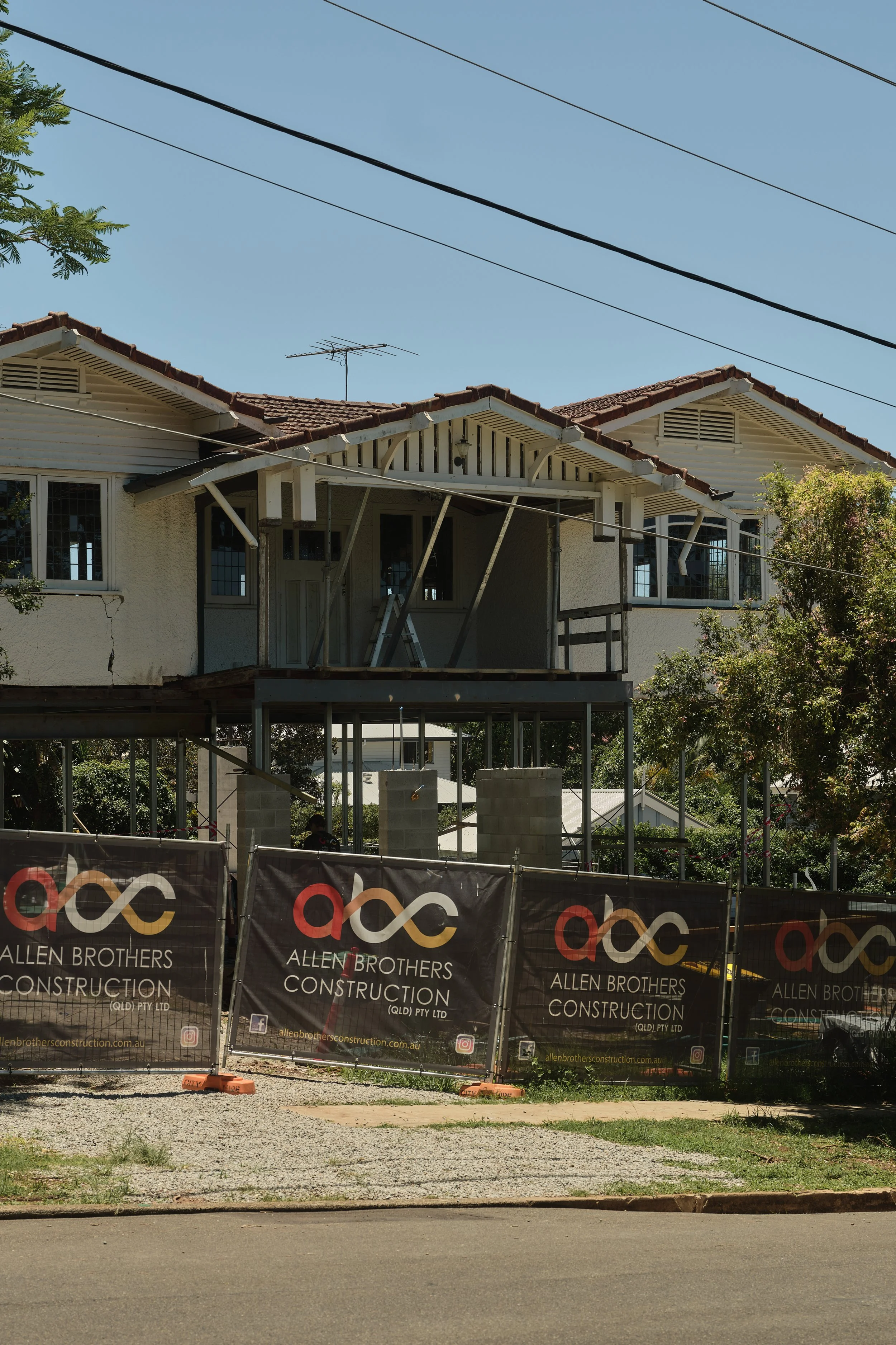 A two‑storey home undergoing a major architectural renovation in Corinda, showing the original upper level retained above new steel framing and concrete block construction. Temporary site fencing displays Allen Brothers Construction branding, with th