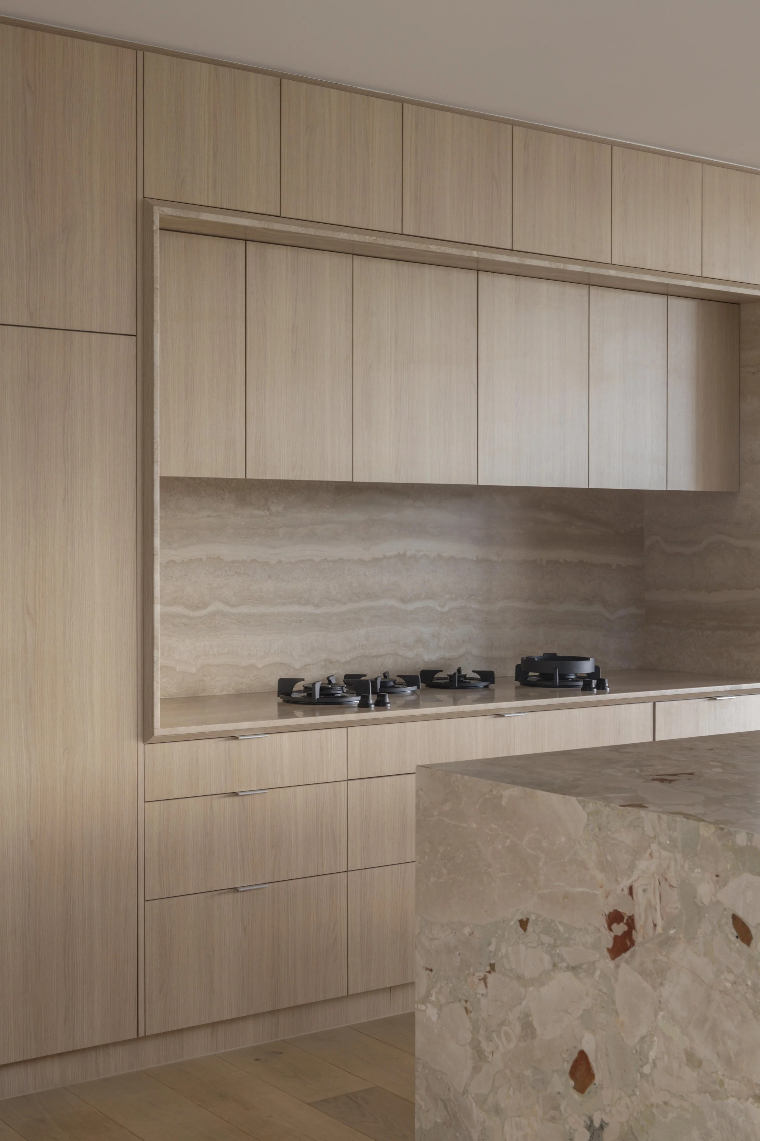 This refined kitchen at Project124 features warm timber cabinetry, a seamless stone benchtop and splashback, and integrated gas cooking set within a minimalist, handle‑free design. The sculptural island bench introduces a striking natural stone with 
