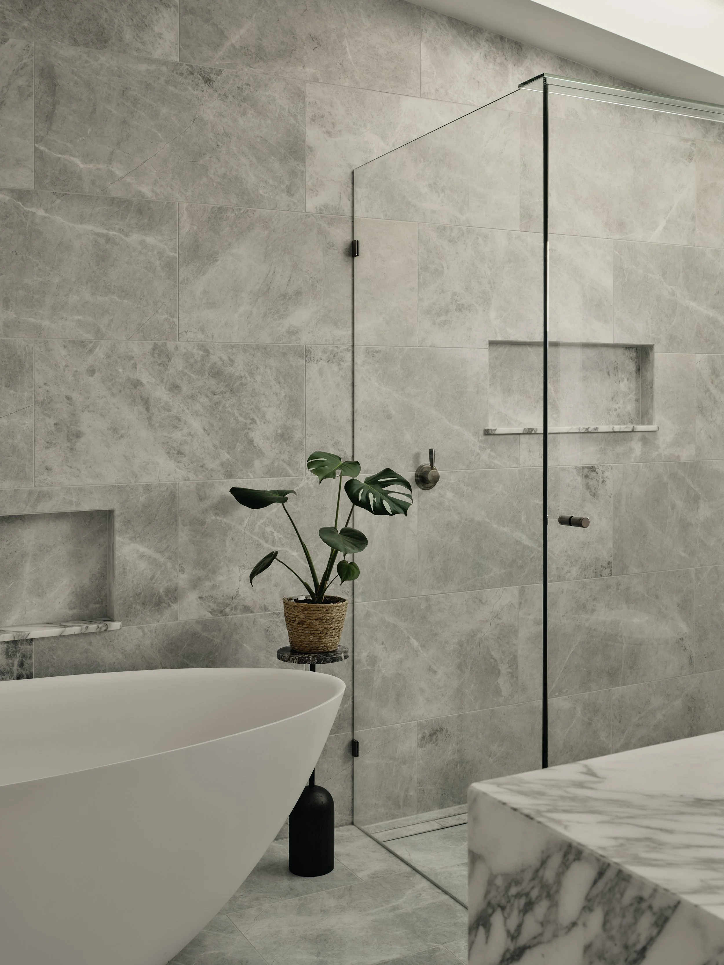 Modern bathroom with a freestanding bathtub, a glass shower enclosure, a potted plant on a small side table, and marble accents.