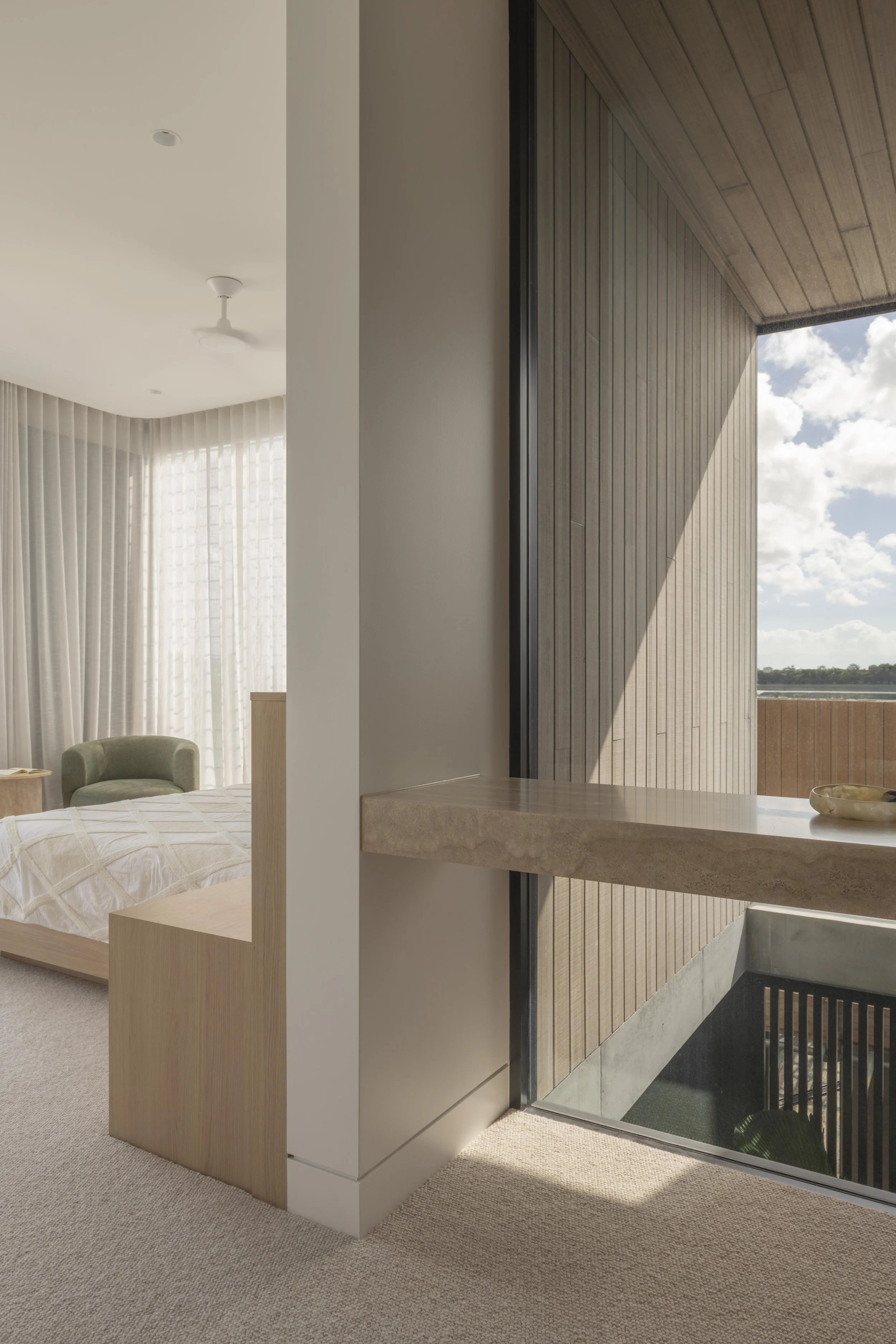 This calming bedroom at Project124 features a soft neutral palette, a geometric‑patterned bedspread, and a sculptural green accent chair that adds subtle colour to the space. Sheer curtains filter natural light across the room, while a partial wall c