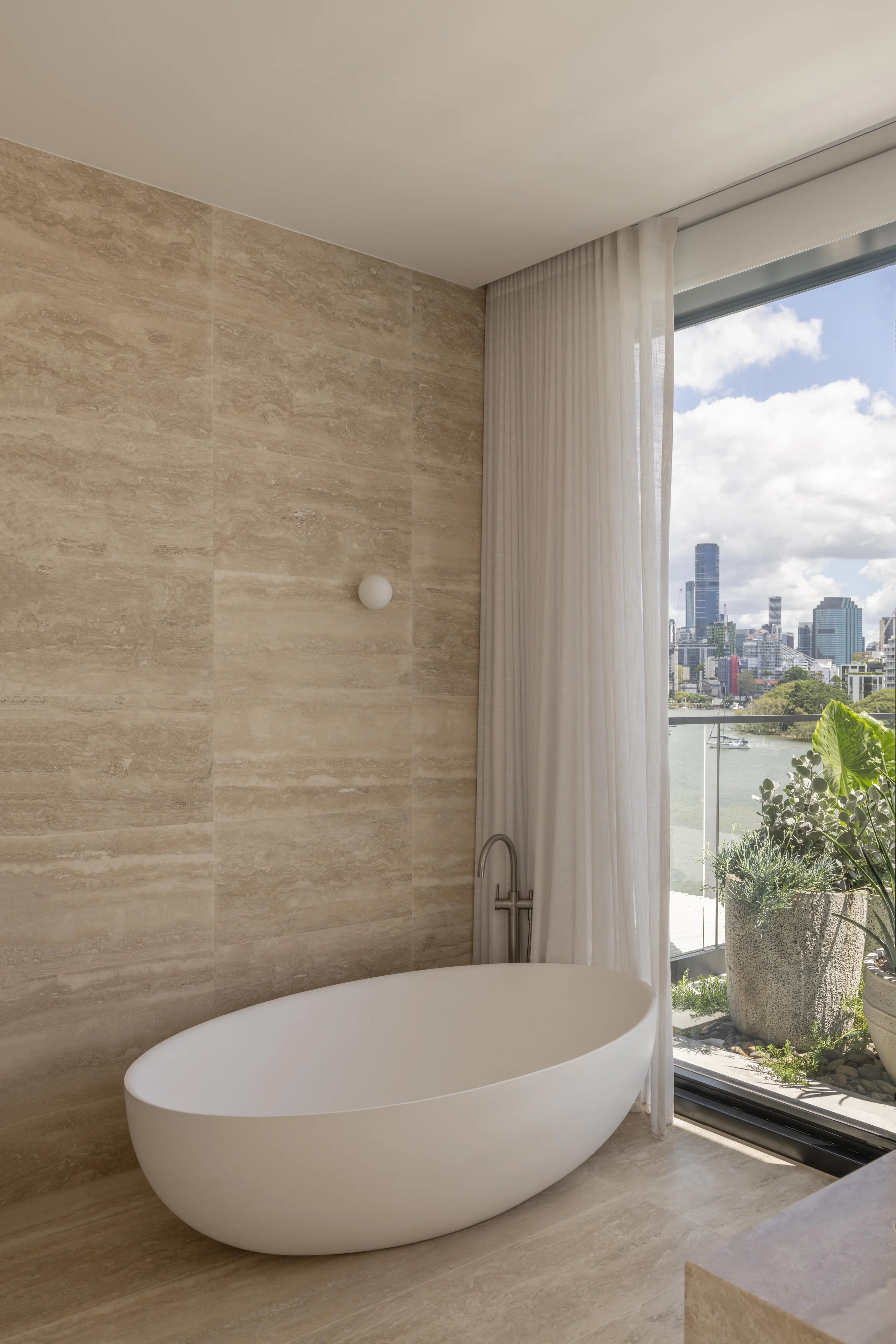 This tranquil bathroom at Project124 features a sculptural freestanding bath set against warm travertine walls, with soft natural light filtering through sheer curtains framing expansive river and city views. The connection to greenery just beyond th