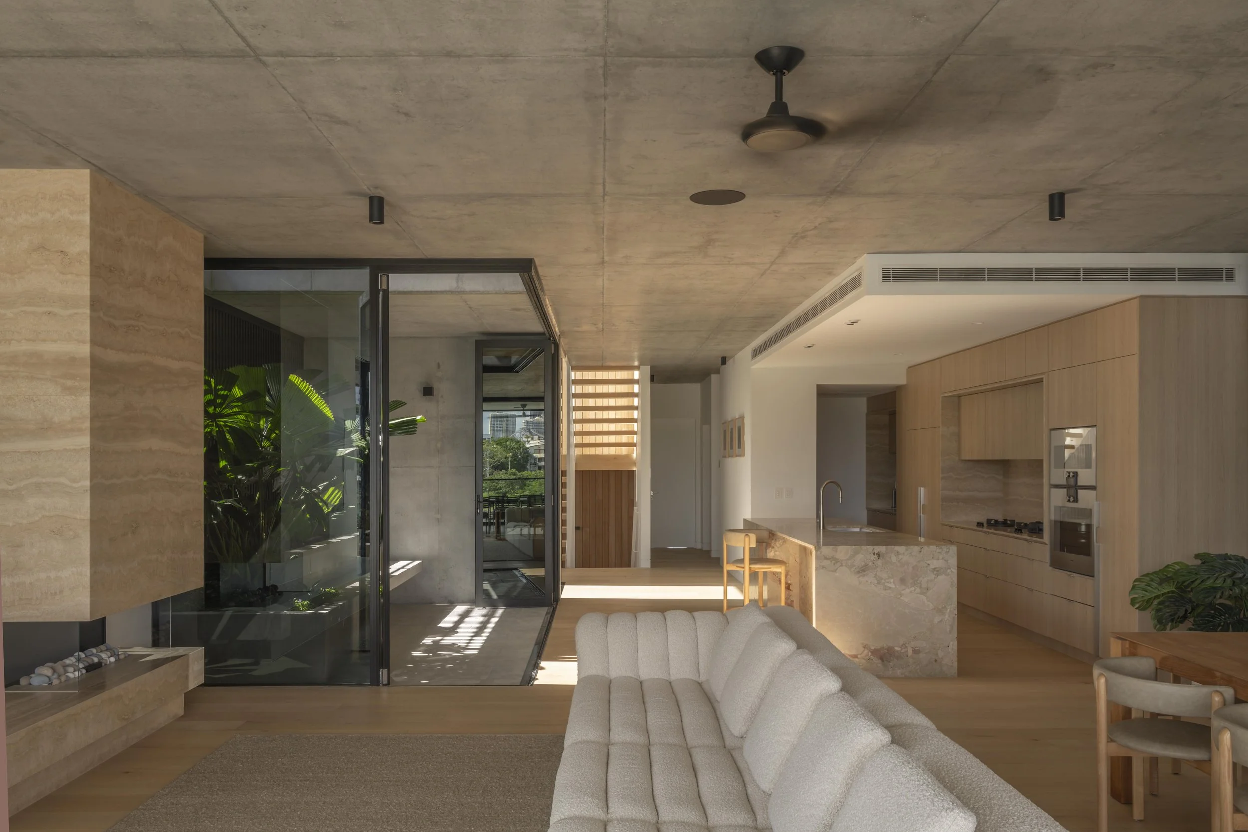 This open‑plan living space at Project124 blends a warm timber kitchen, stone island, and soft, textured furnishings with a lush internal garden framed by full‑height glazing. Natural light washes across the timber floors and off‑form concrete ceilin