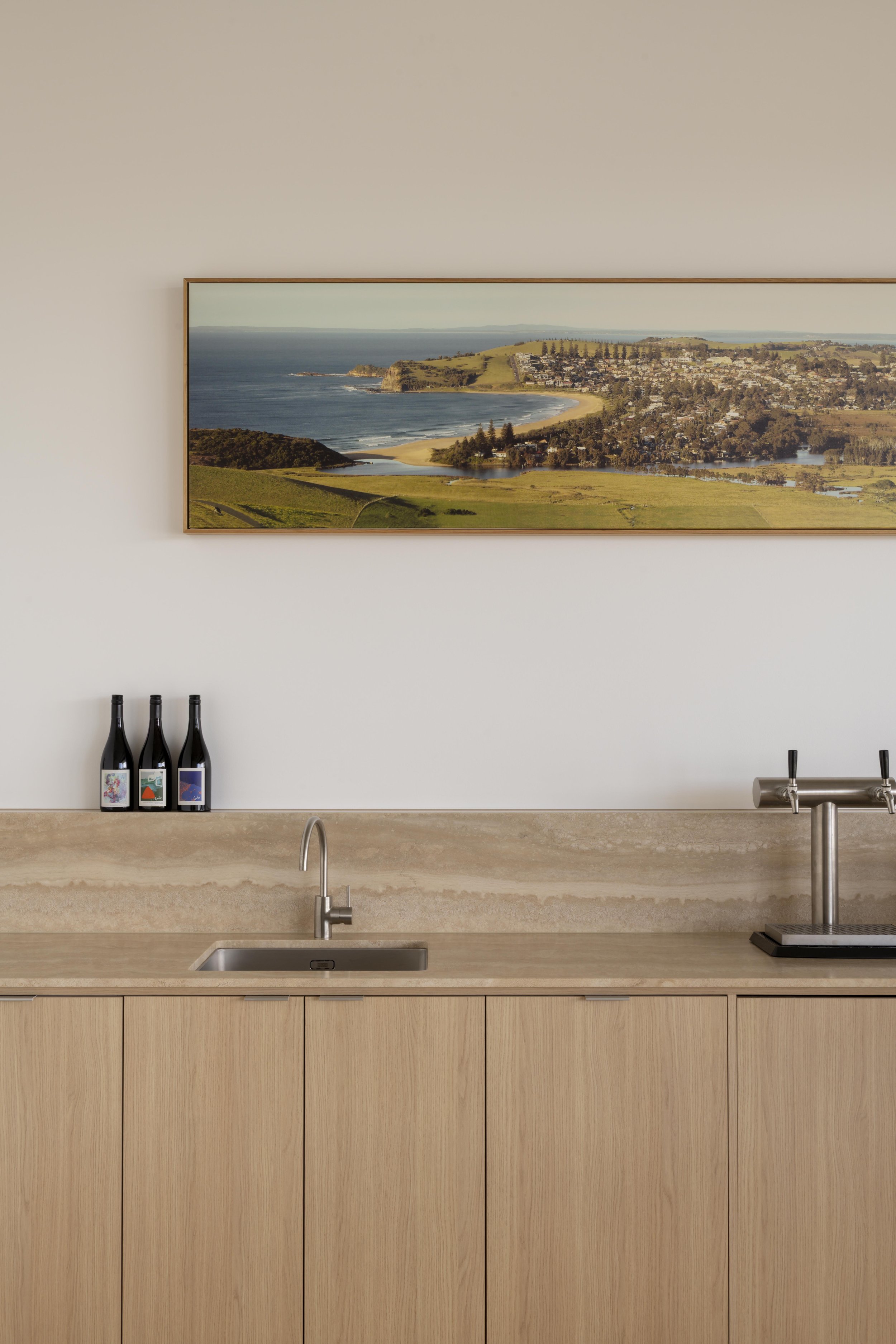 This refined bar zone at Project124 features a stone benchtop with natural veining, warm timber cabinetry, and a panoramic coastal artwork that anchors the space. Integrated wine storage, a stainless‑steel sink, and a dual beer tap elevate the functi