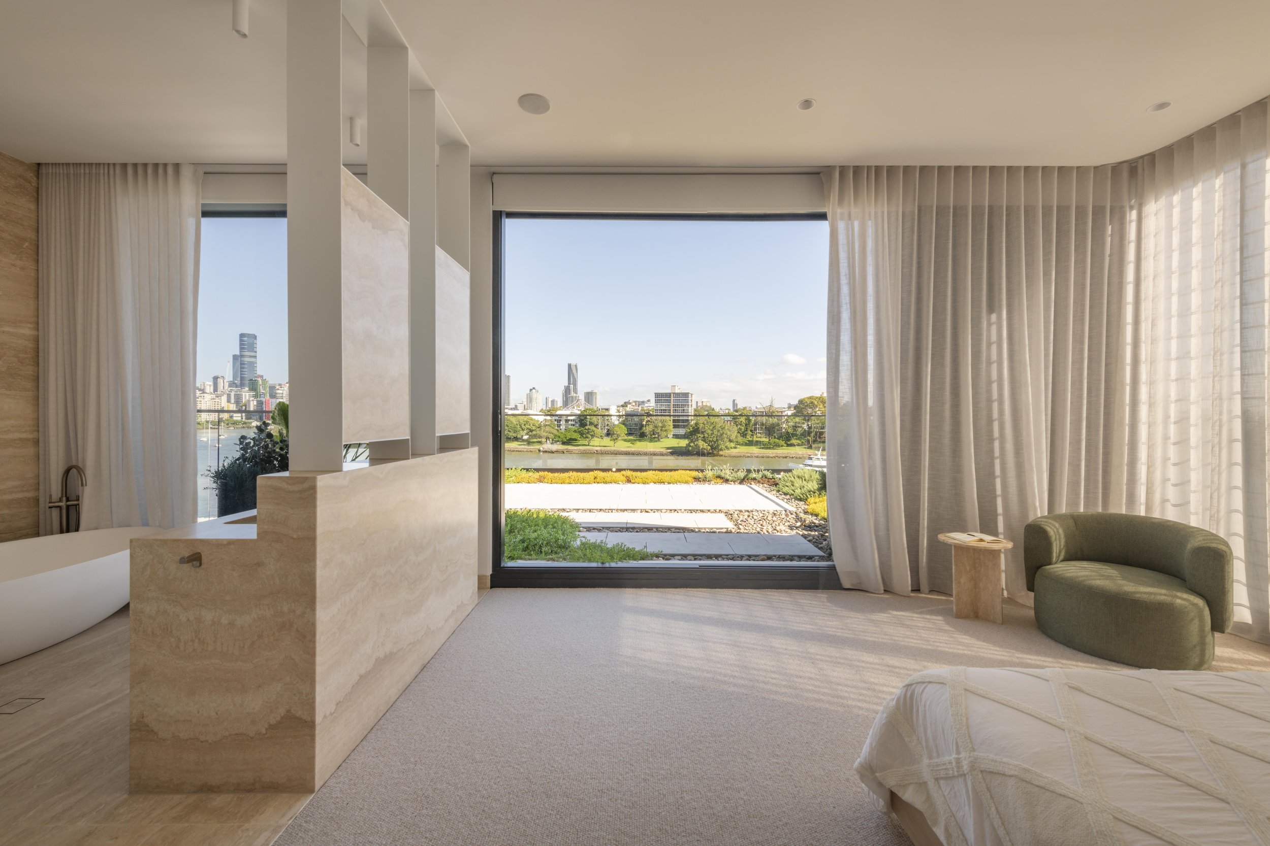 This luxurious bedroom suite at Project124 features a seamless connection to an open ensuite, where a sculptural freestanding bath sits behind a textured stone partition. Floor‑to‑ceiling glazing frames landscaped greenery and distant city views, whi