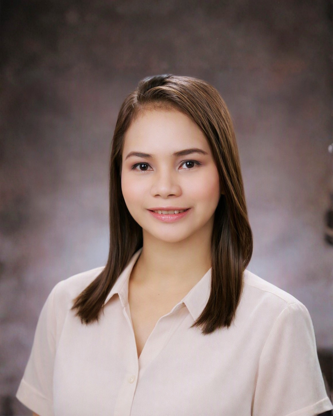 A young woman with shoulder-length brown hair wearing a beige blouse, smiling, against a dark, blurred background.