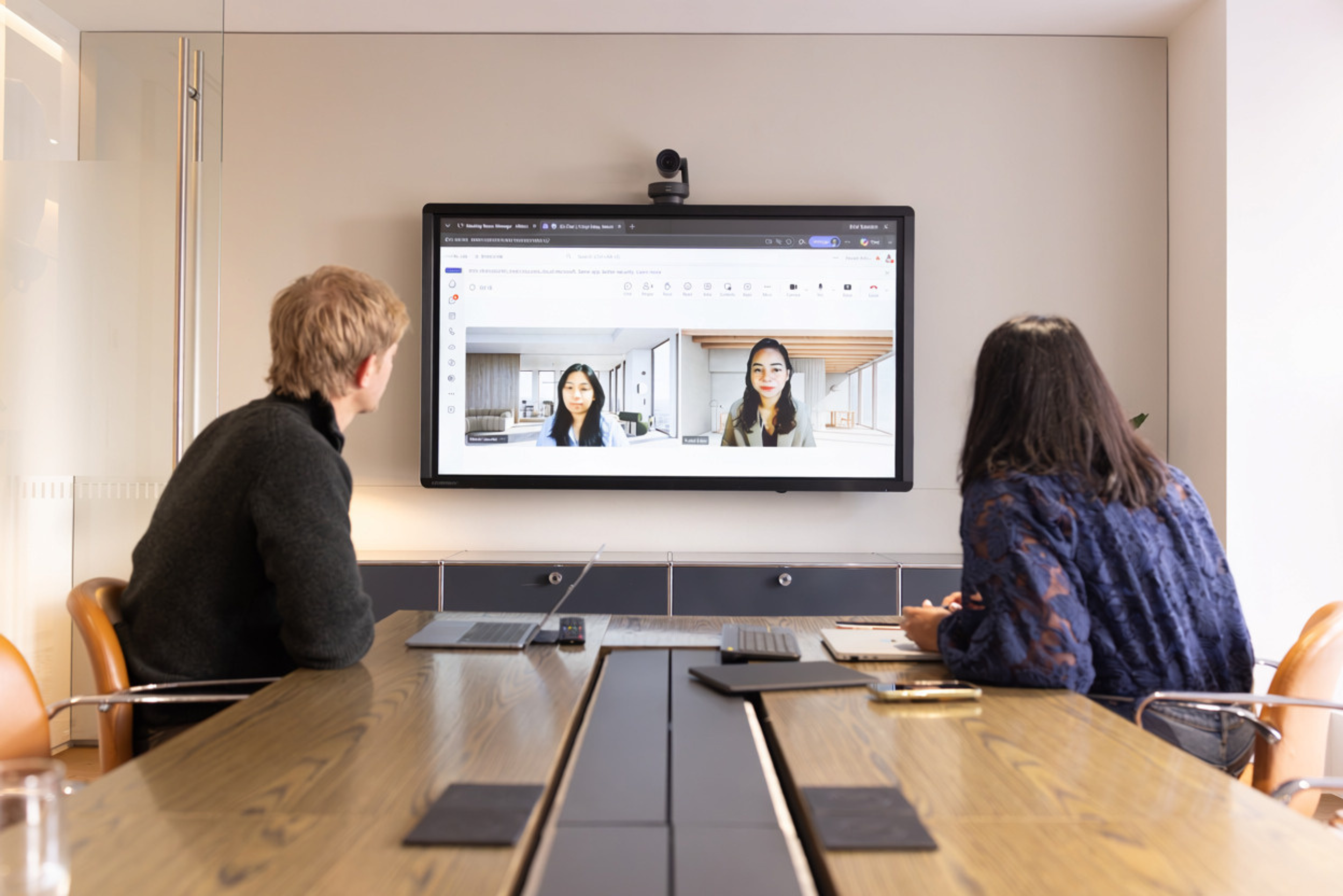 Two people sitting at a conference table, participating in a video call displayed on a large screen, in a modern meeting room.