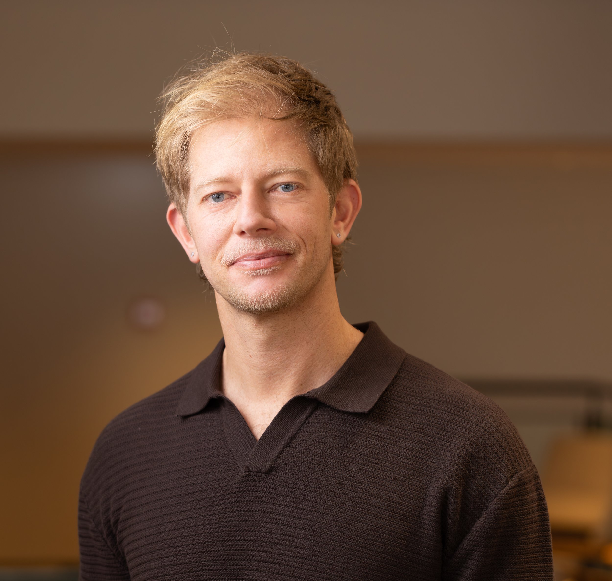 Close-up of a man with blonde hair, blue eyes, earrings, and a slight smile, wearing a dark brown V-neck sweater, standing indoors with a blurred brown background.