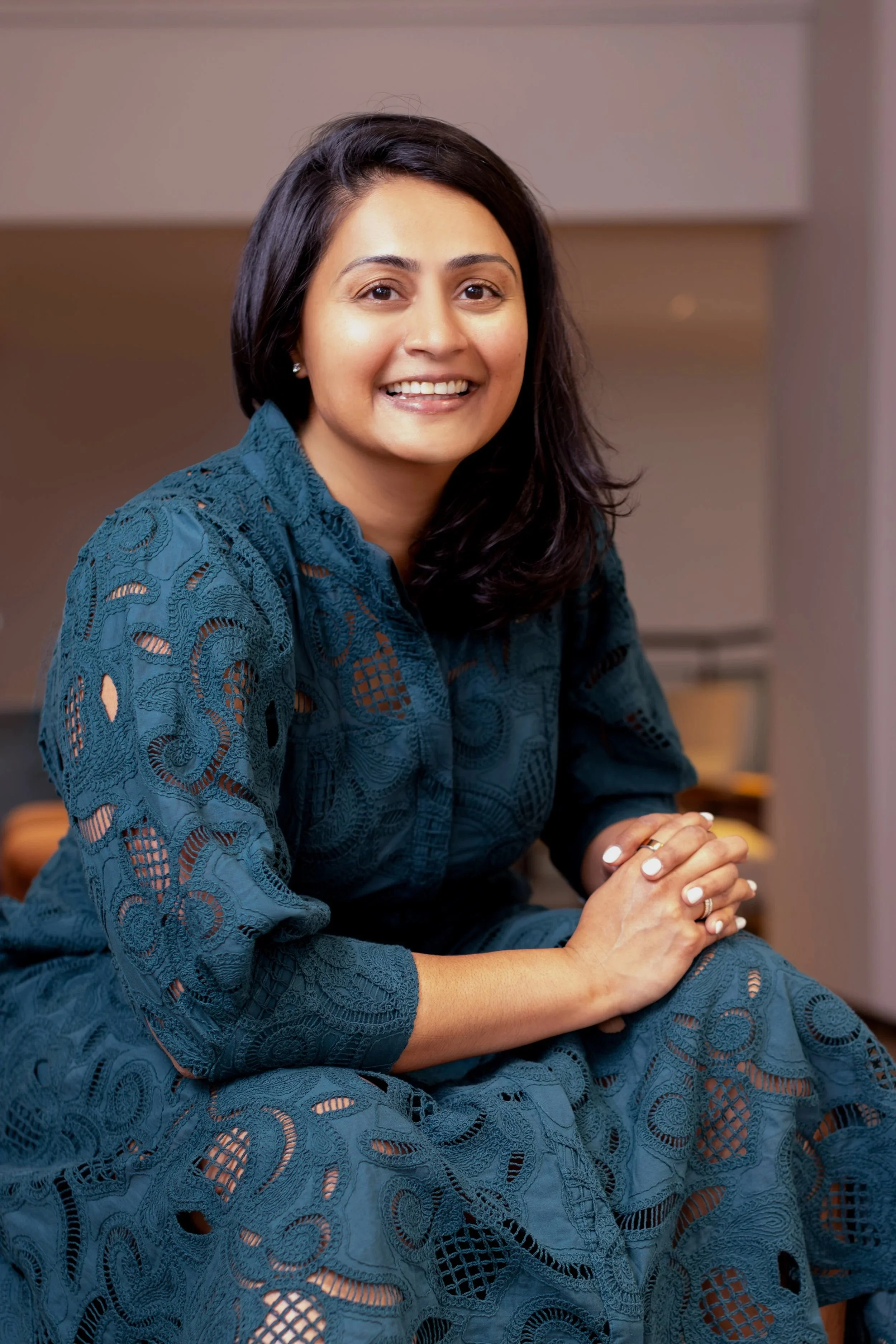 A woman with long dark hair, wearing a teal lace dress, smiling and sitting in an indoor setting.