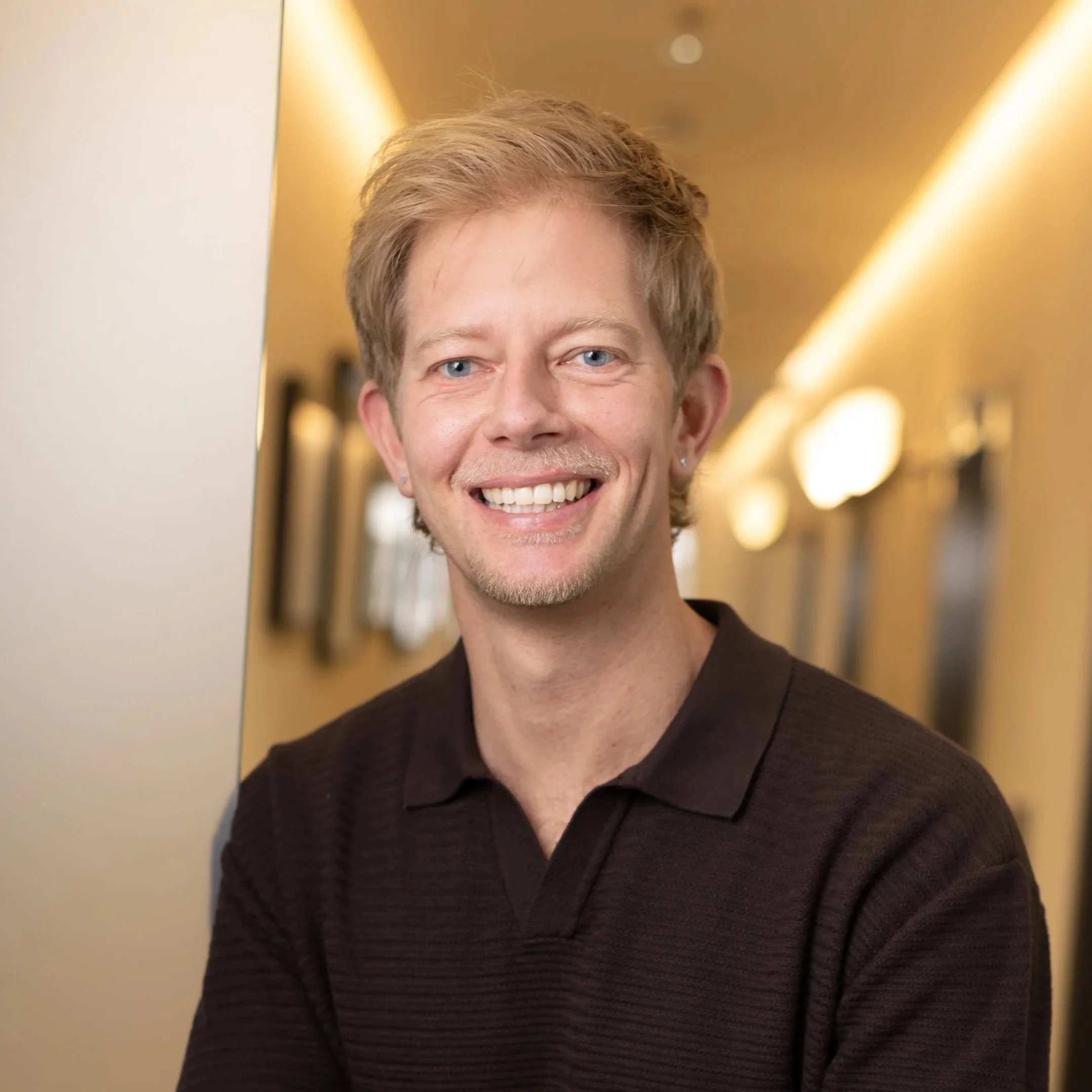 Close-up of a smiling person with short, light brown or blonde hair, blue eyes, and a small earring, wearing a dark ribbed long-sleeve shirt. The background is softly lit with warm yellow tones.