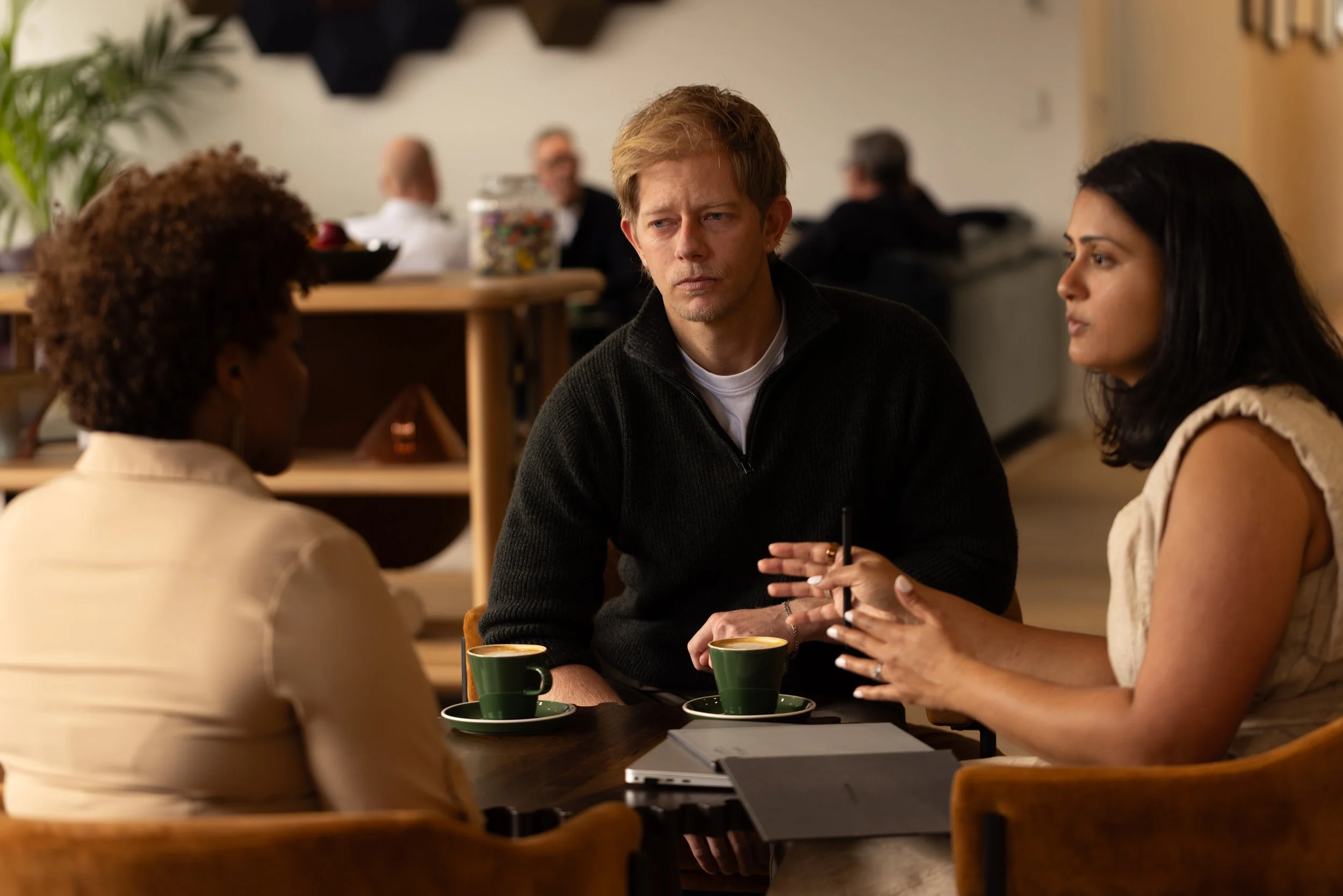 Three people engaged in a serious conversation at a cafe. Two women and one man are sitting around a table with cups of coffee, with other customers blurred in the background.