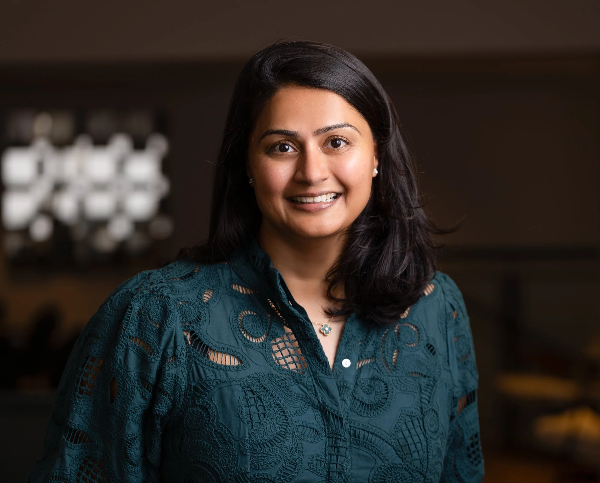 A woman with dark hair and brown eyes wearing a teal lace blouse and a flower-shaped necklace smiling indoors.