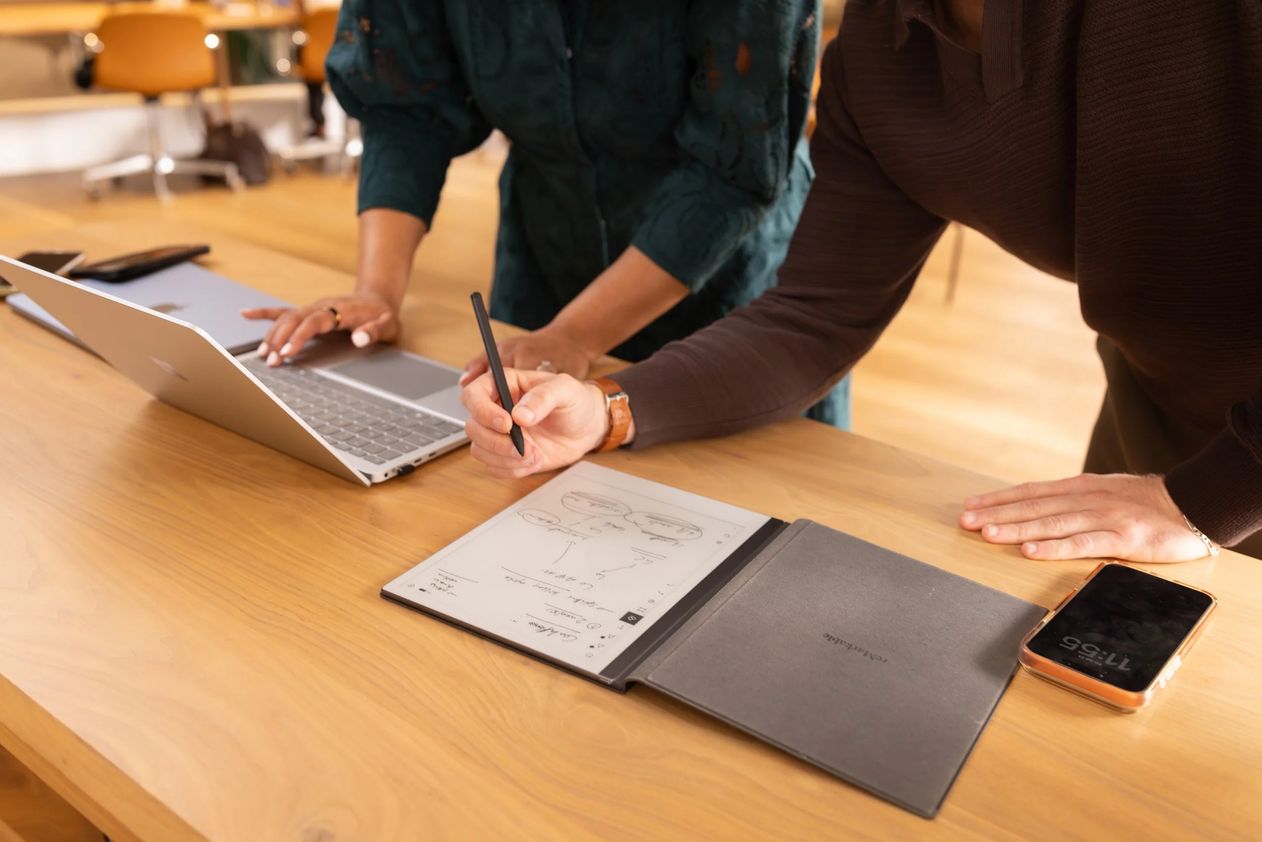 Two people are standing at a wooden table, collaborating on a project with a laptop, tablet, notebook, and smartphone visible.