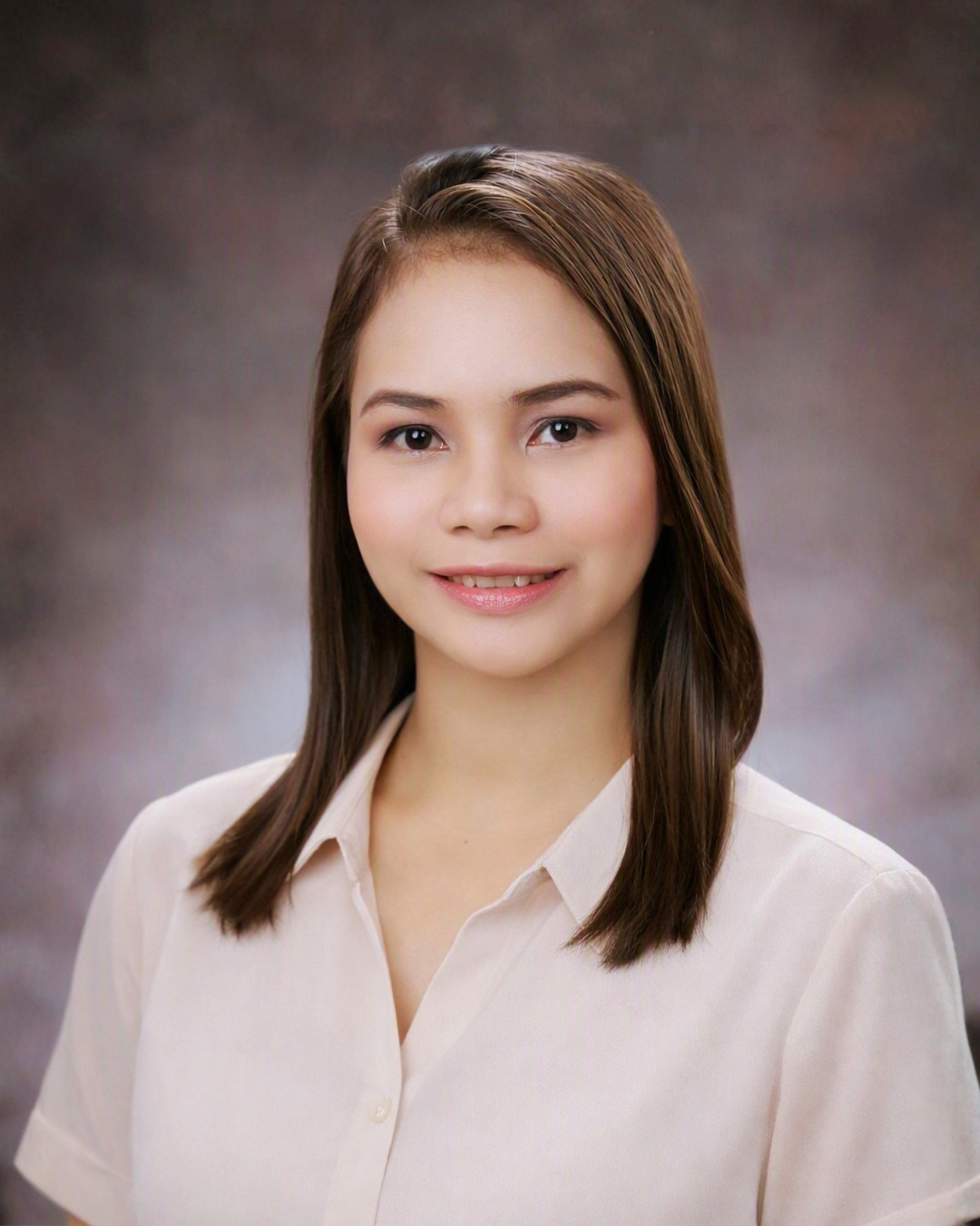 A young woman with straight brown hair, wearing a light-colored button-up shirt, smiling softly against a blurred dark background.