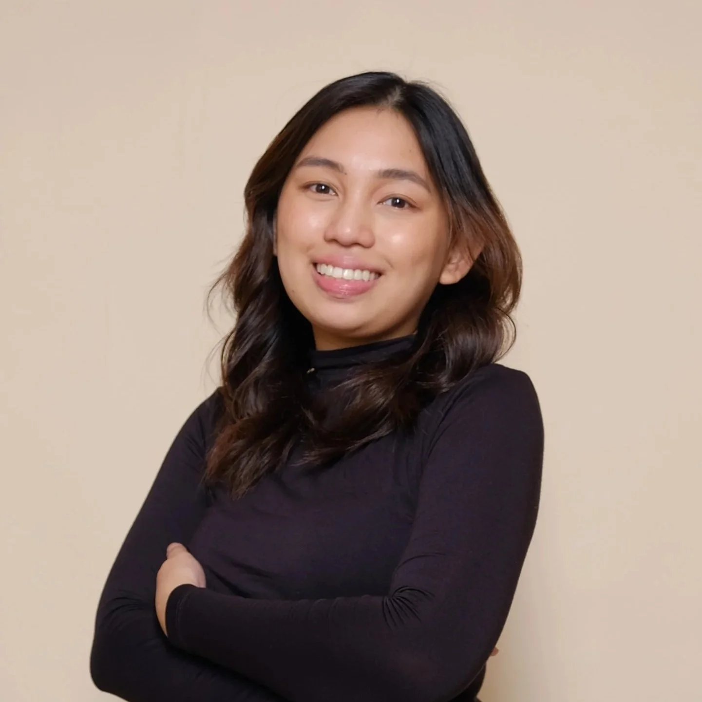 A woman with shoulder-length dark hair smiling with arms crossed, wearing a black long-sleeve top, standing against a plain beige background.