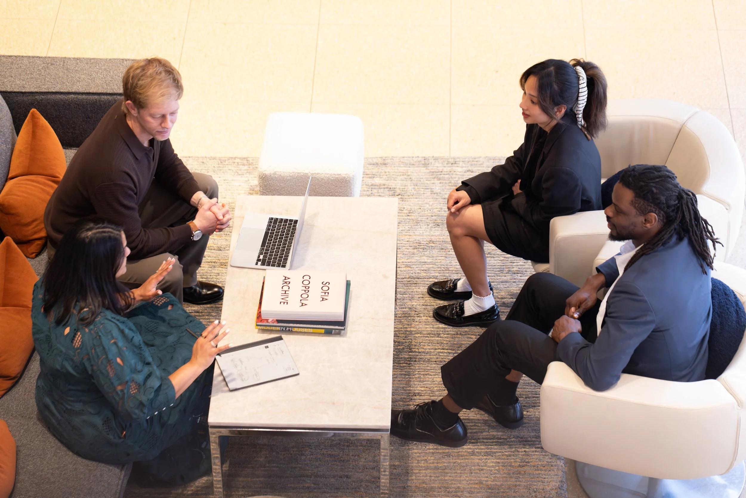 A group of diverse people having a meeting around a white table with books, notebooks, and a laptop inside a modern office.