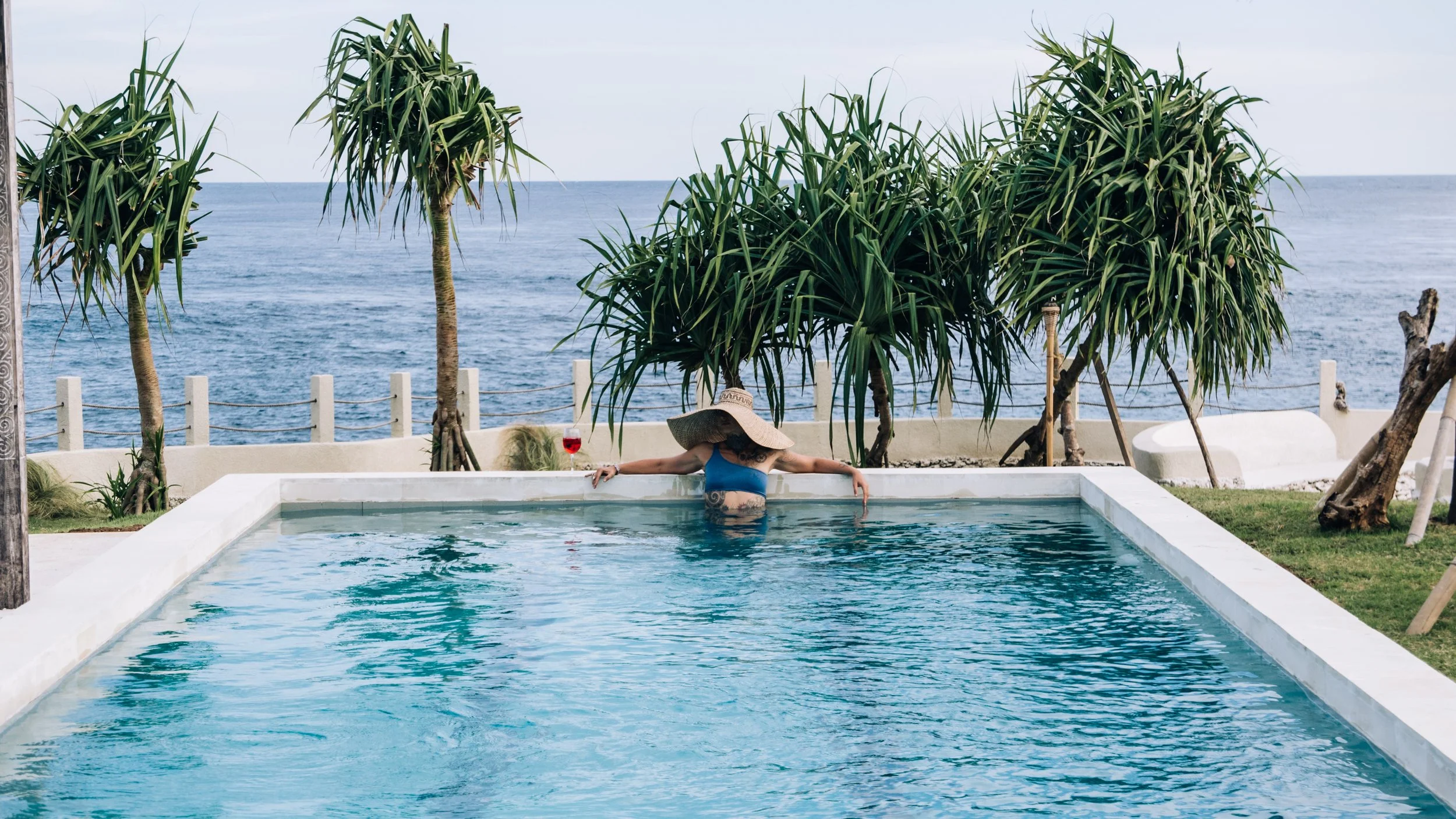 A woman in a blue swimsuit and wide-brimmed hat relaxing in a swimming pool by the ocean, with a glass of red wine nearby and tropical trees in the background.