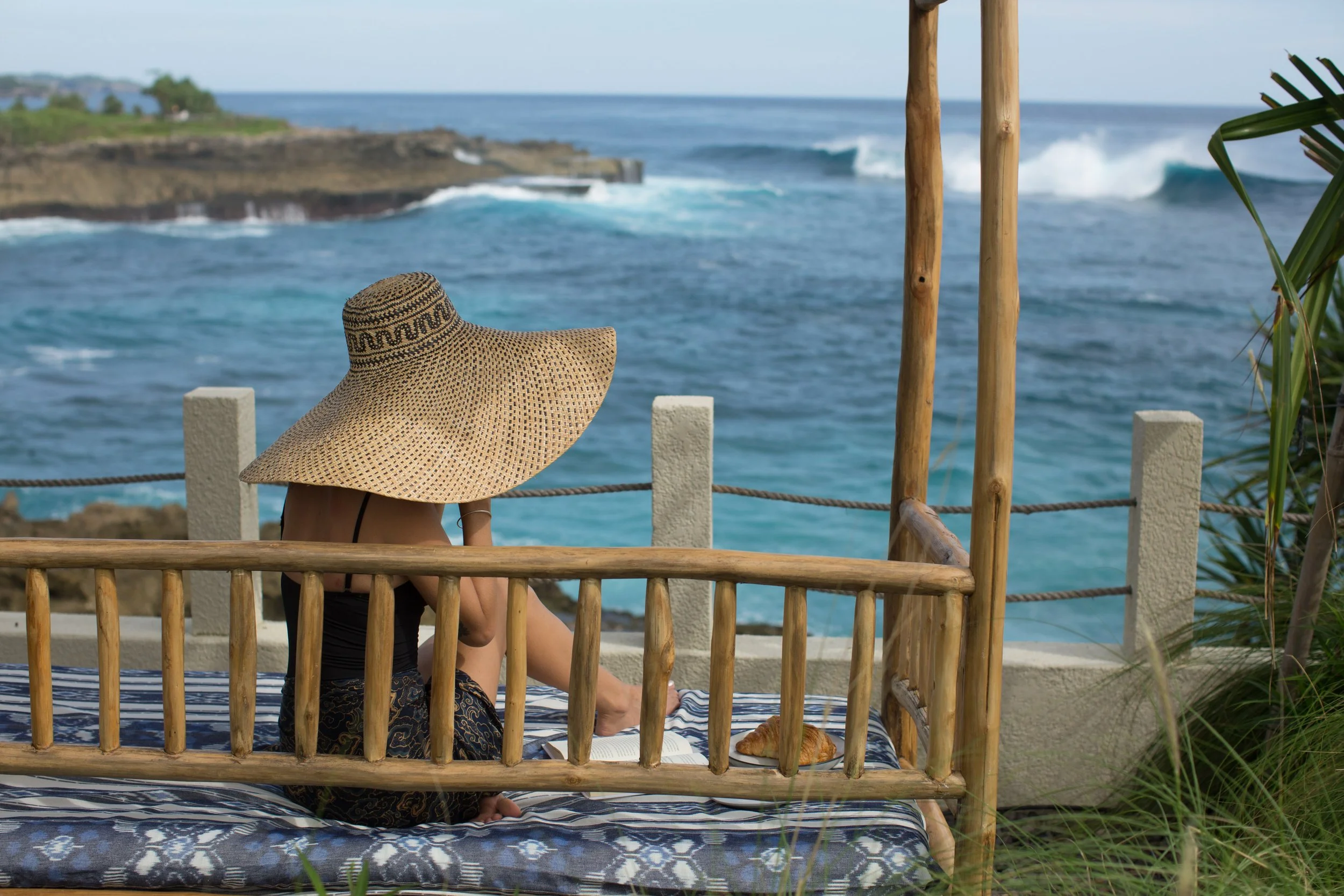 A person sitting on a bamboo bed wearing a large straw hat, looking at the ocean with waves crashing against rocks in the background.