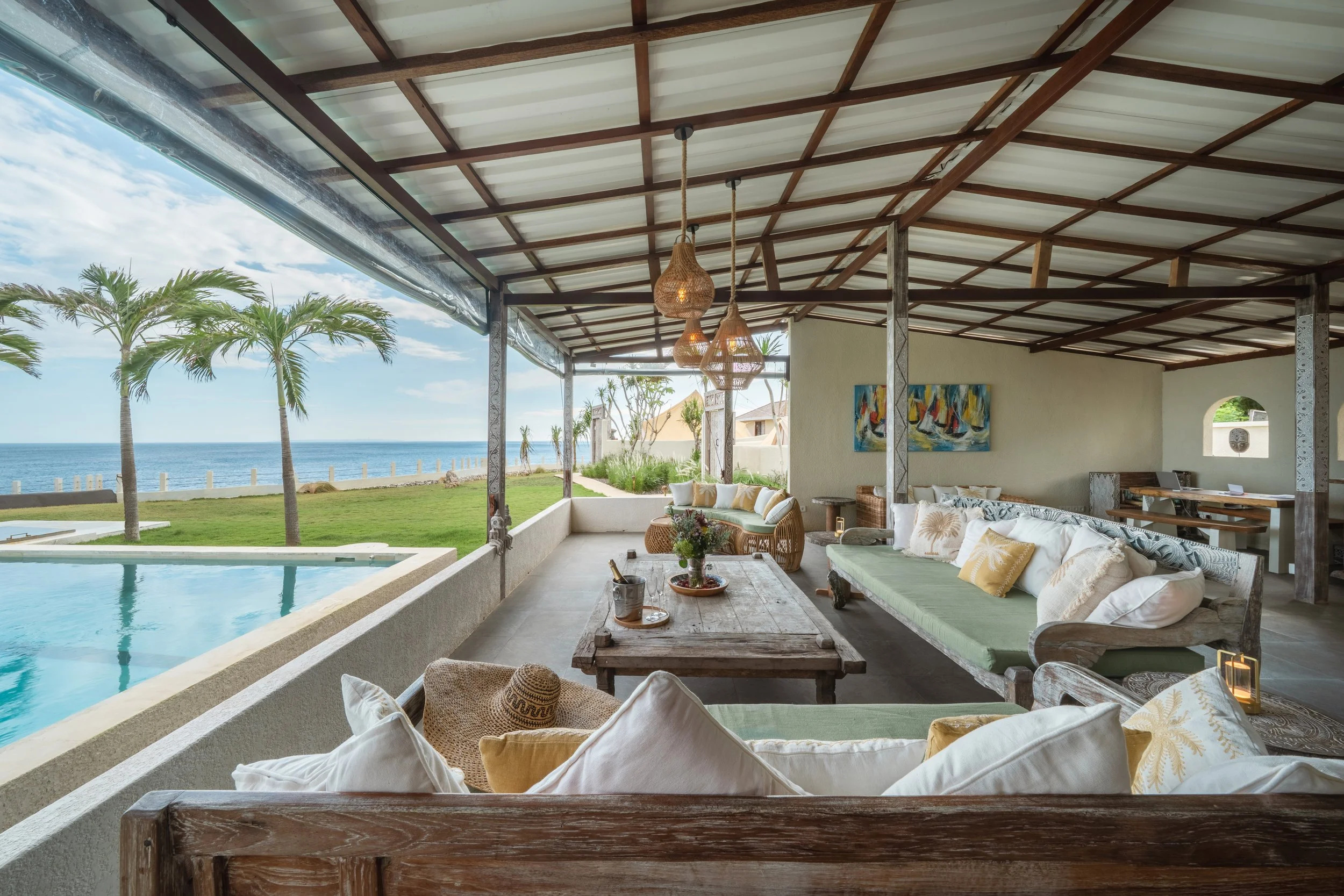 Beachside covered outdoor lounge with wicker sofas and a wooden coffee table, overlooking a grassy area, palm trees, and the ocean.