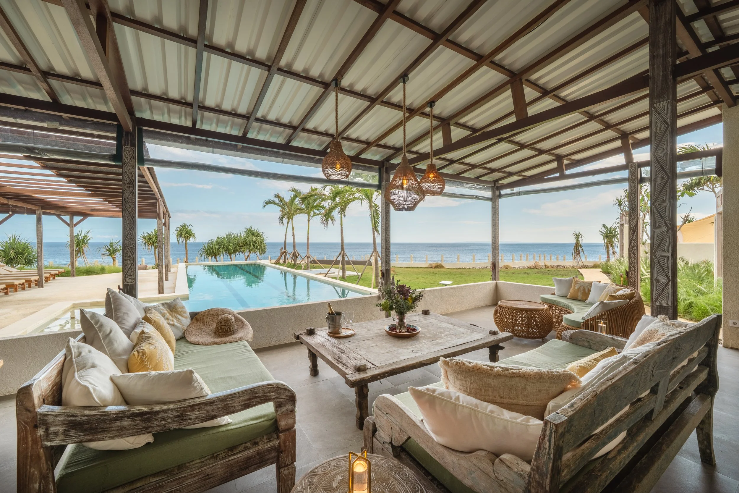 Open-air beachside lounge area with wooden furniture, cushions, a rustic coffee table, and a view of a swimming pool, palm trees, and the ocean.