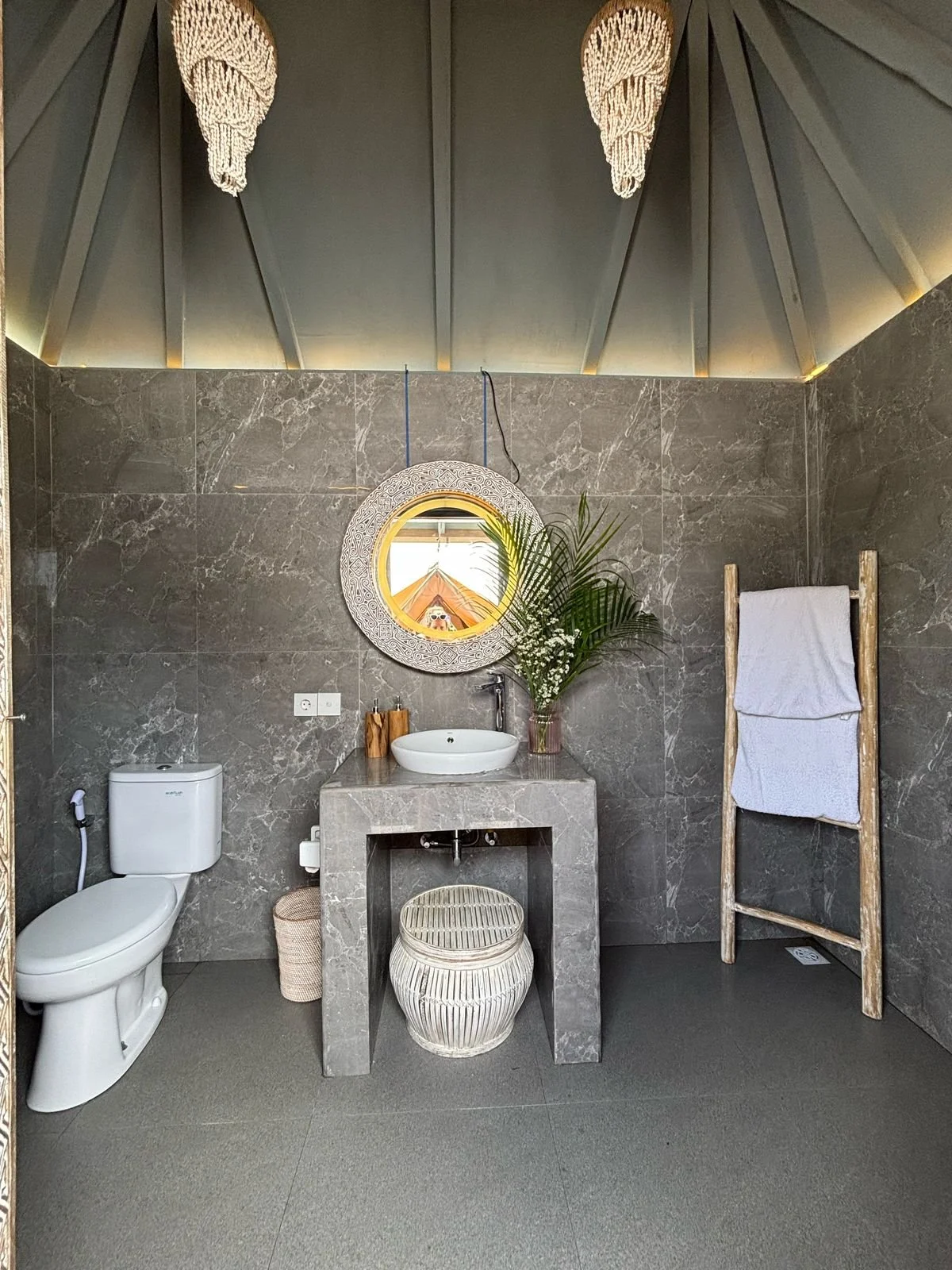 A modern bathroom with dark gray marble walls, a white toilet, a concrete sink with a round mirror above it, a tall wooden towel rack with white towels, and a small wicker stool underneath the sink, with a decorative fern plant and two wooden contain