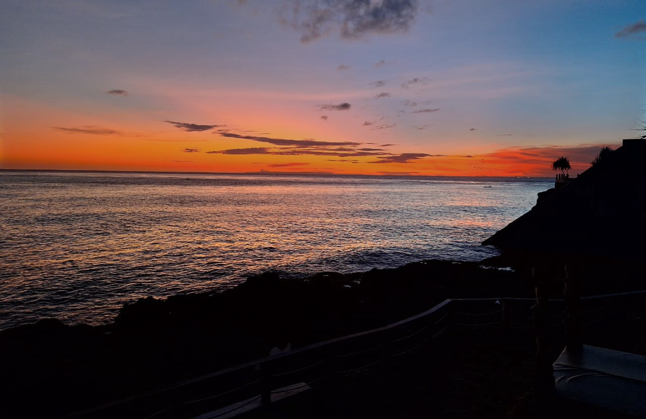 Sunset over the ocean with colorful sky, silhouette of a rocky shoreline and a small house with palm trees on a hillside.