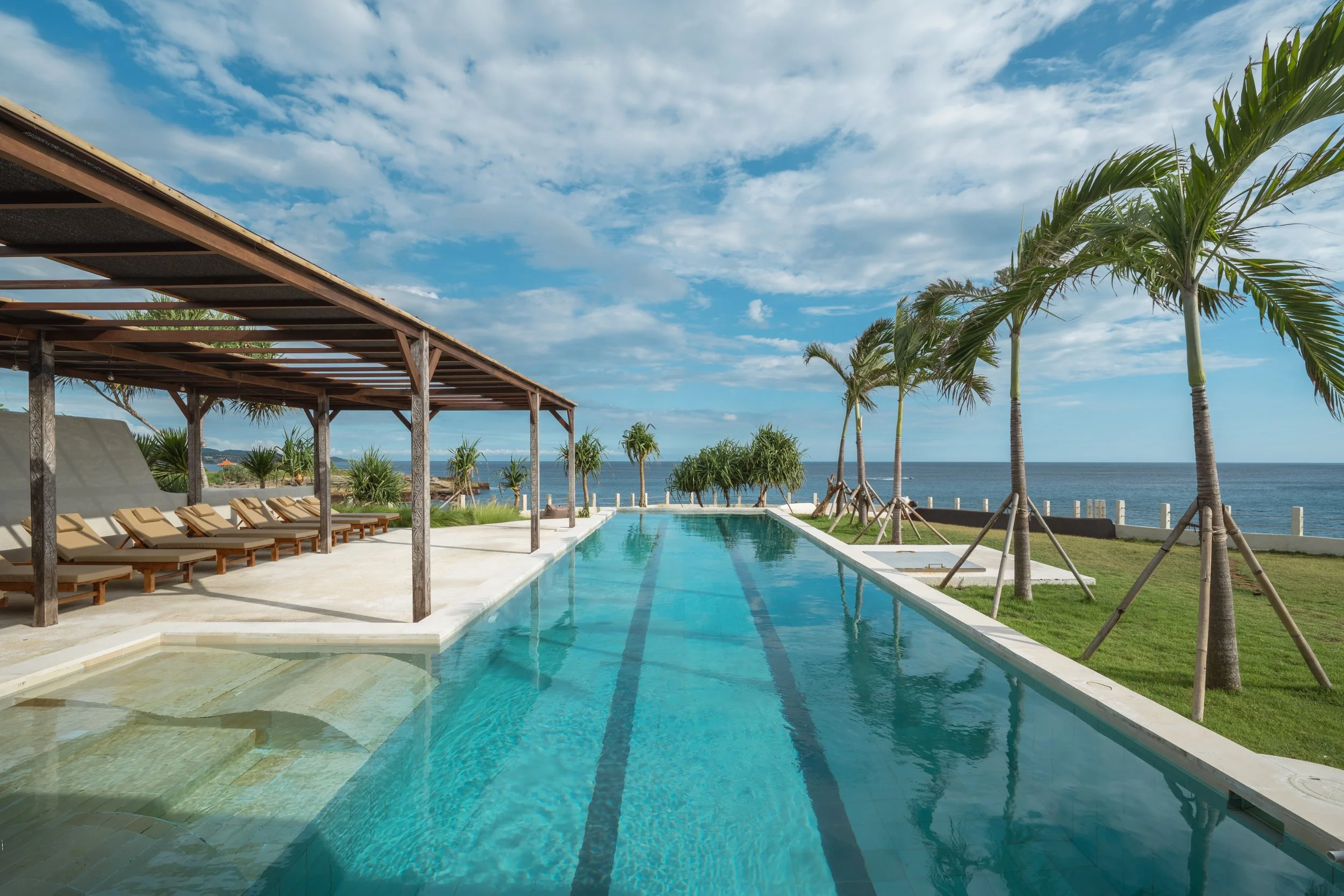 A luxurious outdoor swimming pool area overlooking the ocean, with a row of lounge chairs under a wooden pergola on the left and palm trees on the right. The sky is partly cloudy with a bright, sunny atmosphere.