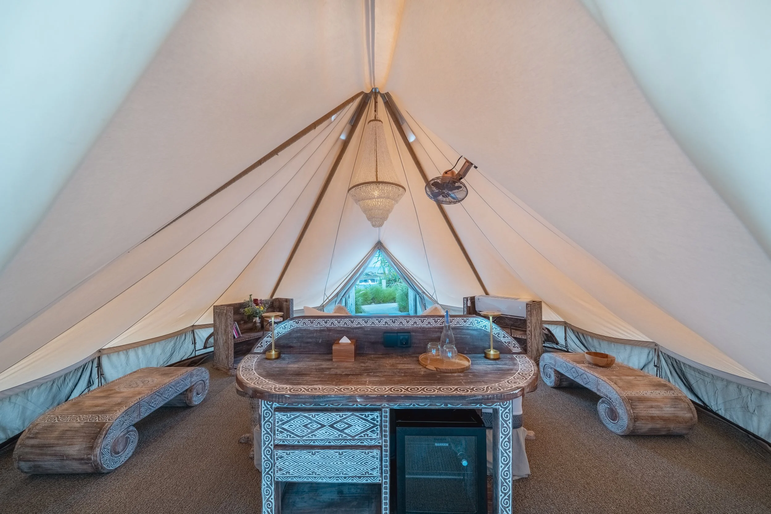 Interior of a spacious glamping tent with a wooden table, decorative benches, a chandelier, and a small window showing greenery outside.