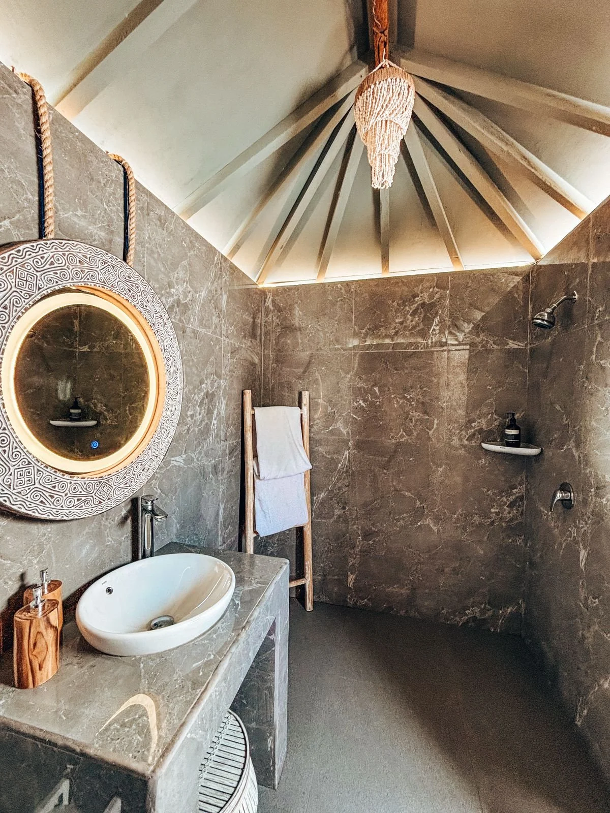 A bathroom with brown marble walls, a white oval sink on a marble countertop, a round mirror with intricate design, a towel ladder with white towels, and a shower area with two shelves and a showerhead.