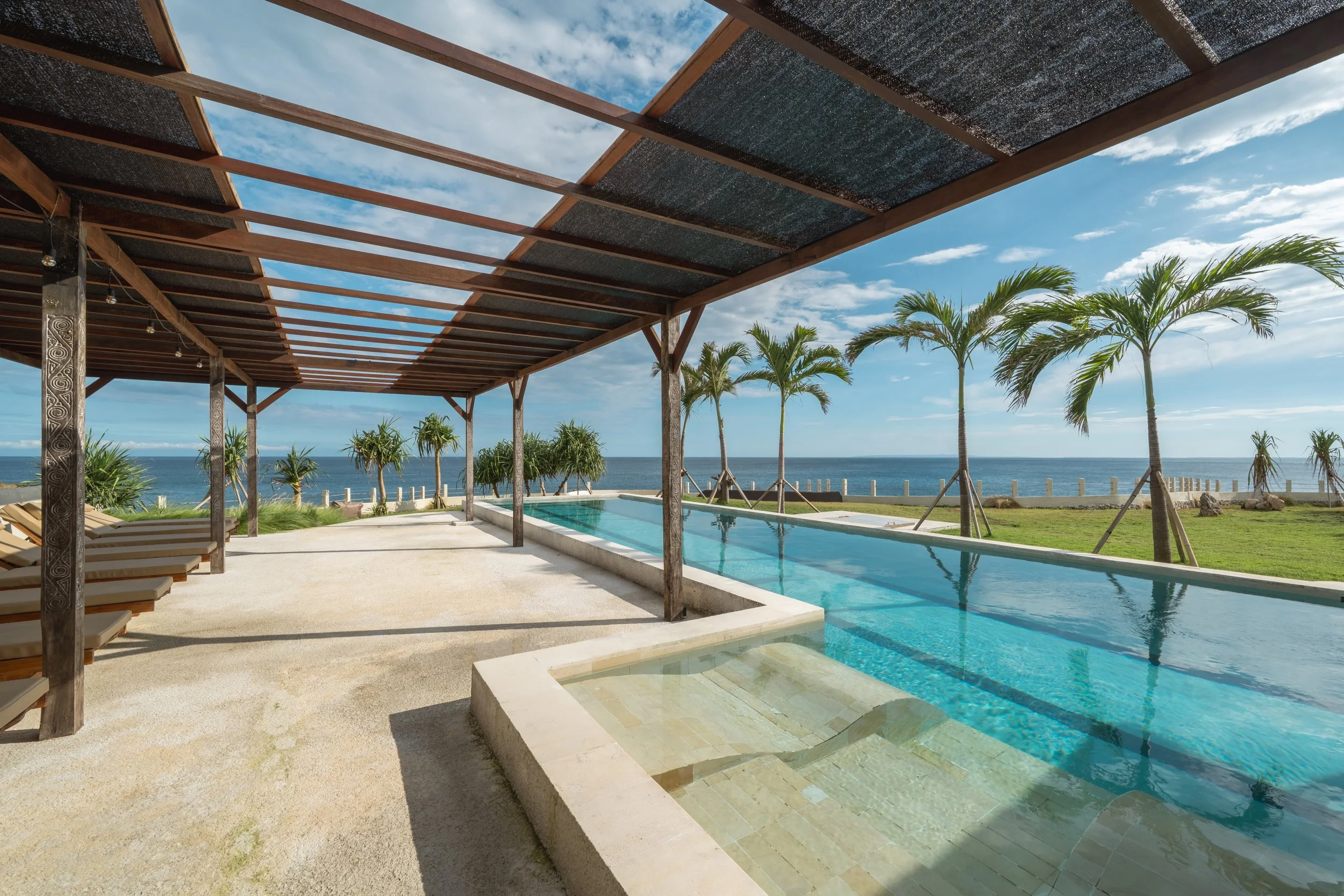 A view of a poolside area with a pergola, lounge chairs, and palm trees overlooking the ocean on a sunny day.