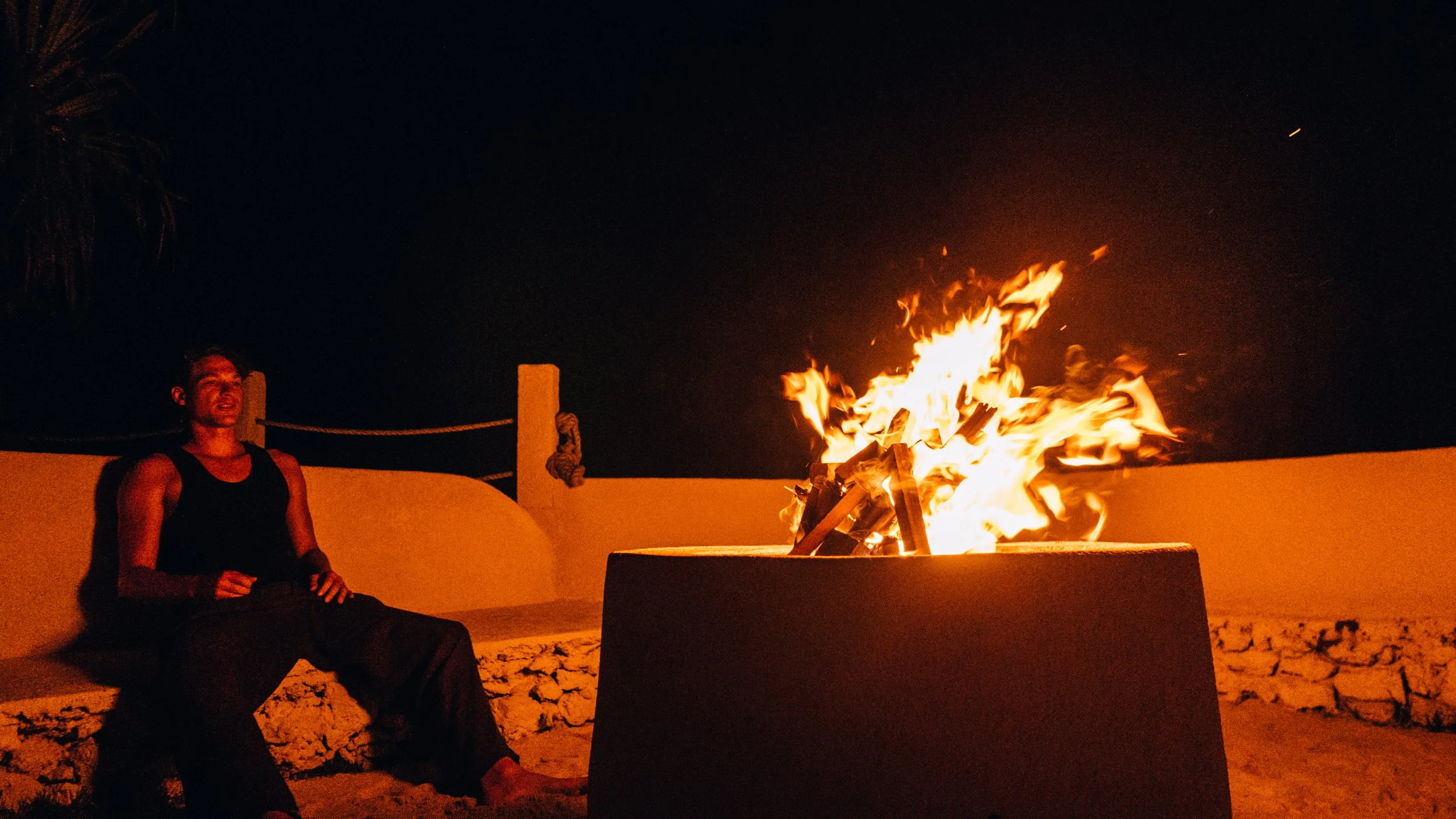 A person sitting on a beach at night near a large bonfire.
