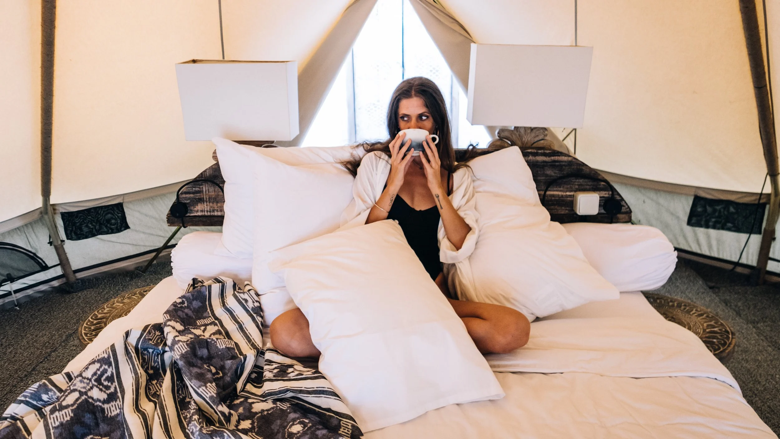 Woman sitting cross-legged on a bed inside a tent, holding a mug to her mouth, with multiple pillows around her.