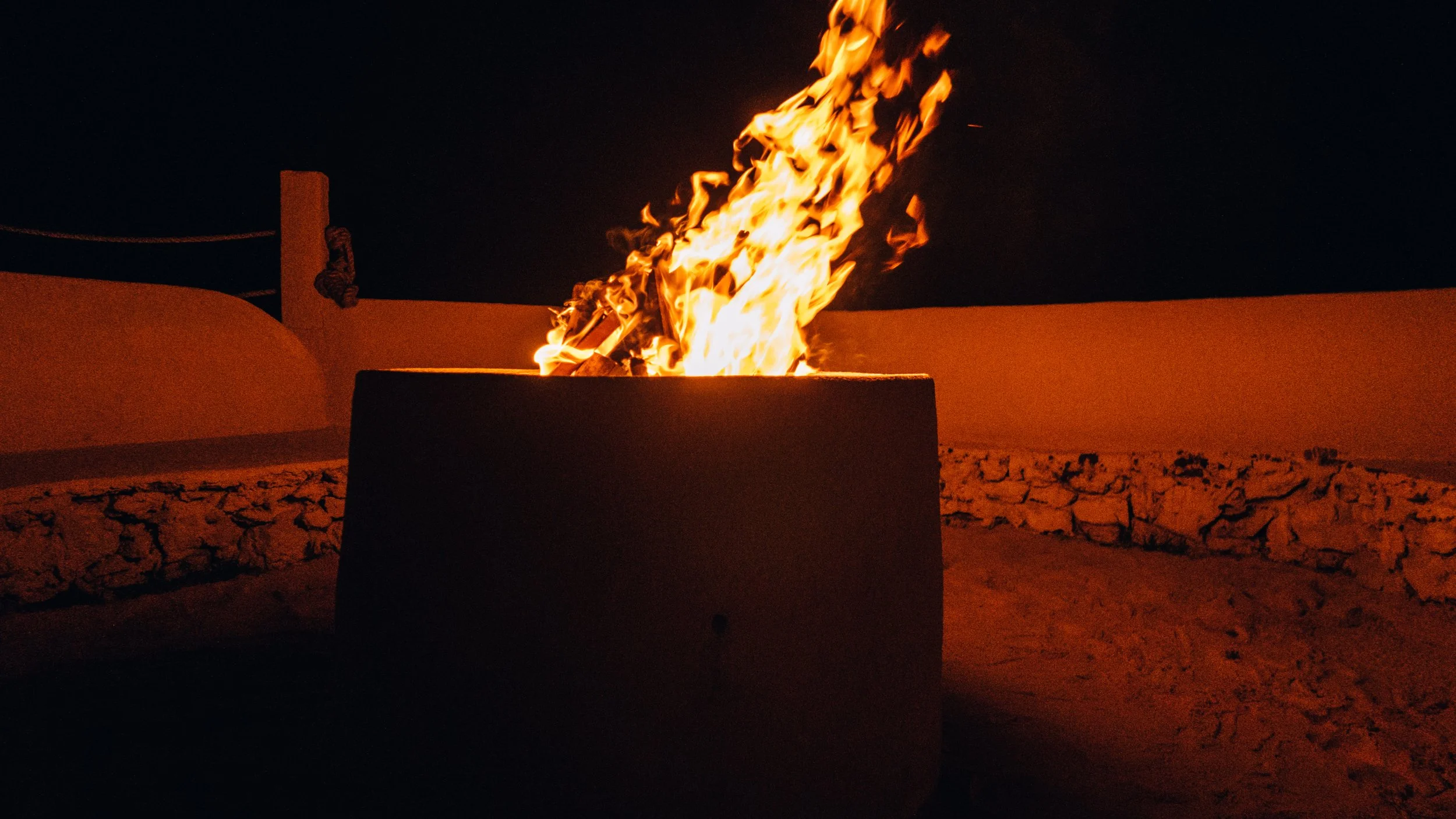 A fire burning in a fire pit at night, surrounded by a low stone wall with a clear dark sky in the background.