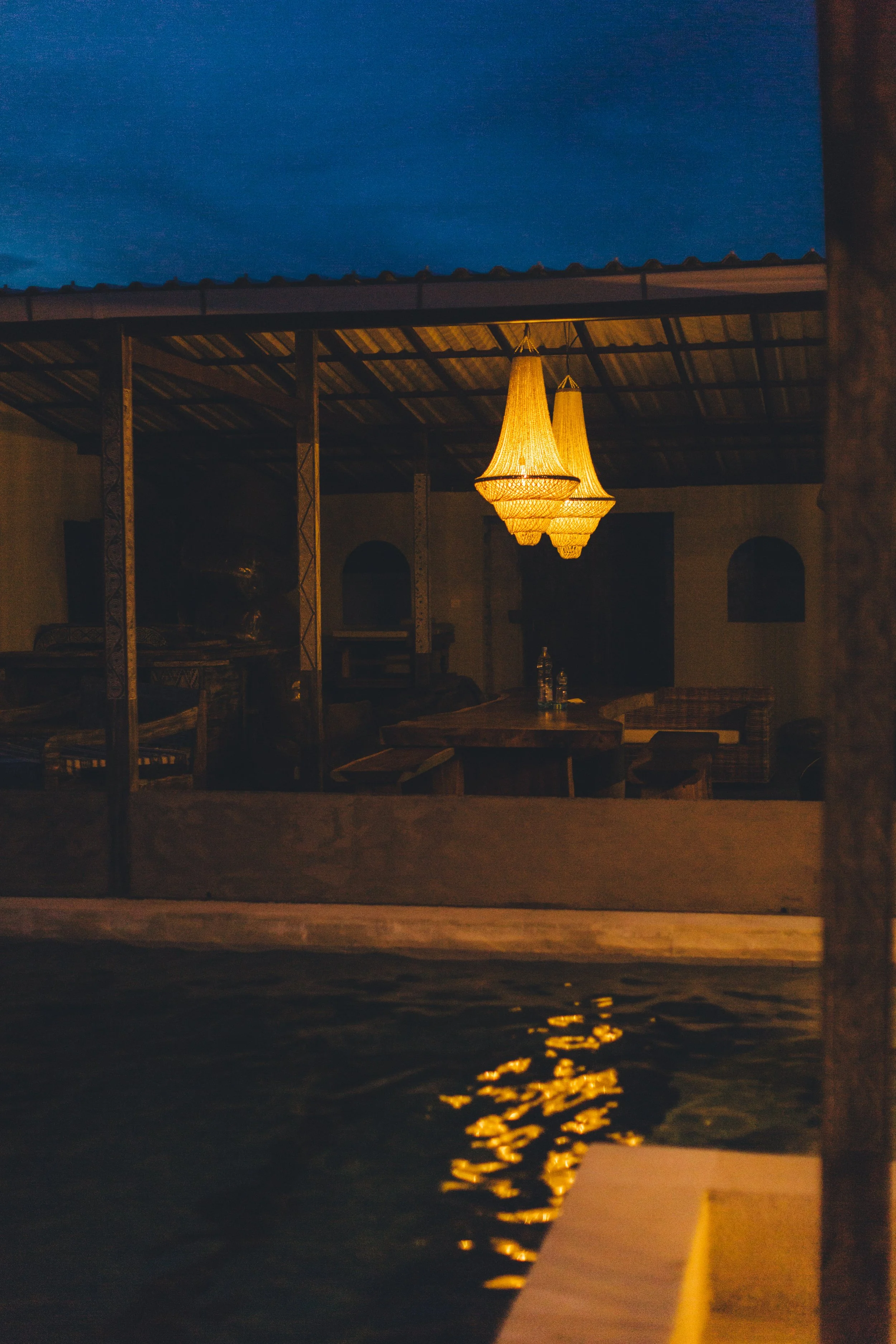 Indoor area with warm lighting chandeliers hanging over tables, visible from outside through a window reflecting water, at night.