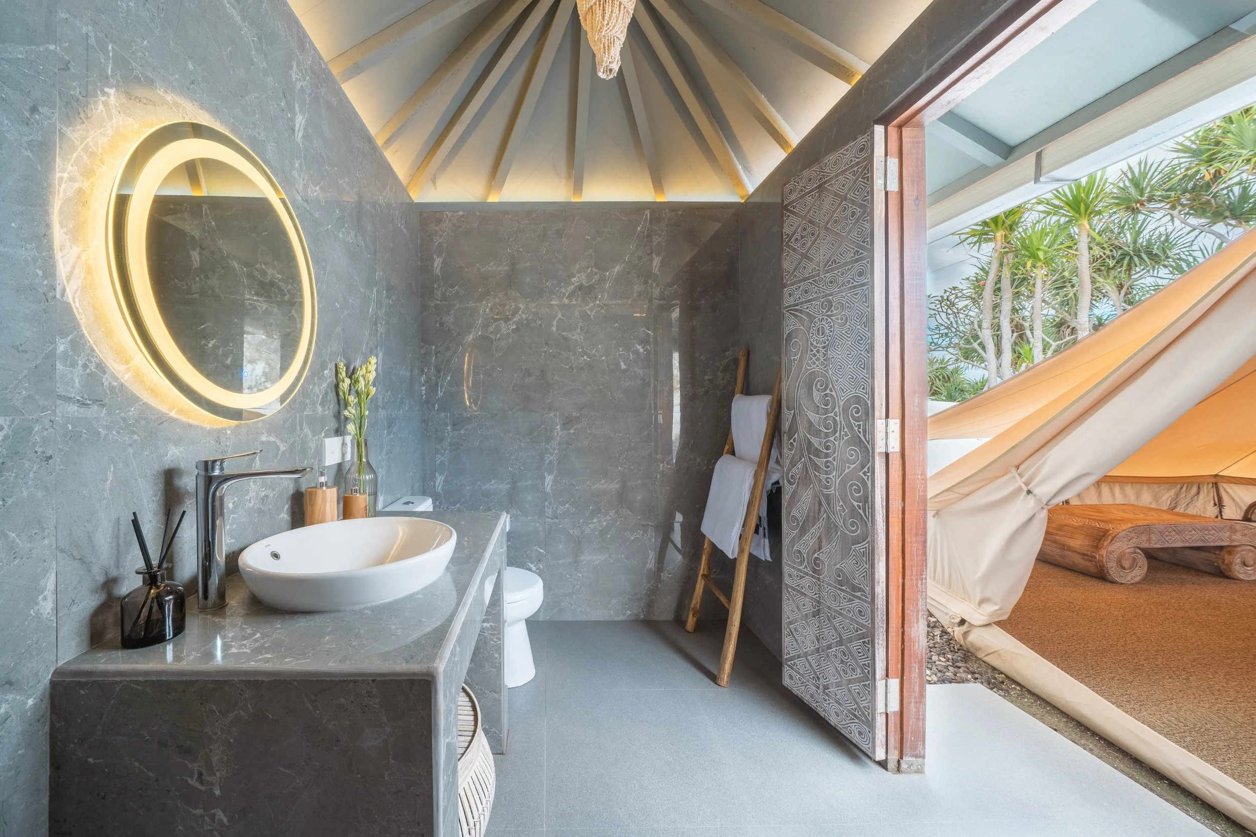 Bathroom with gray marble walls and countertop, round illuminated mirror, white vessel sink, green plant in a glass bottle, towels on a wooden ladder, open door leading to outdoor tent with trees and a wooden lounge