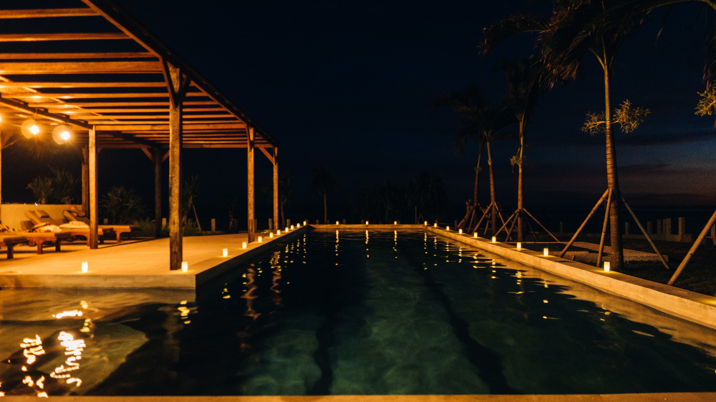 Nighttime view of a luxurious pool surrounded by candles, palm trees, and a wooden pergola with lounge chairs on a beachside property.