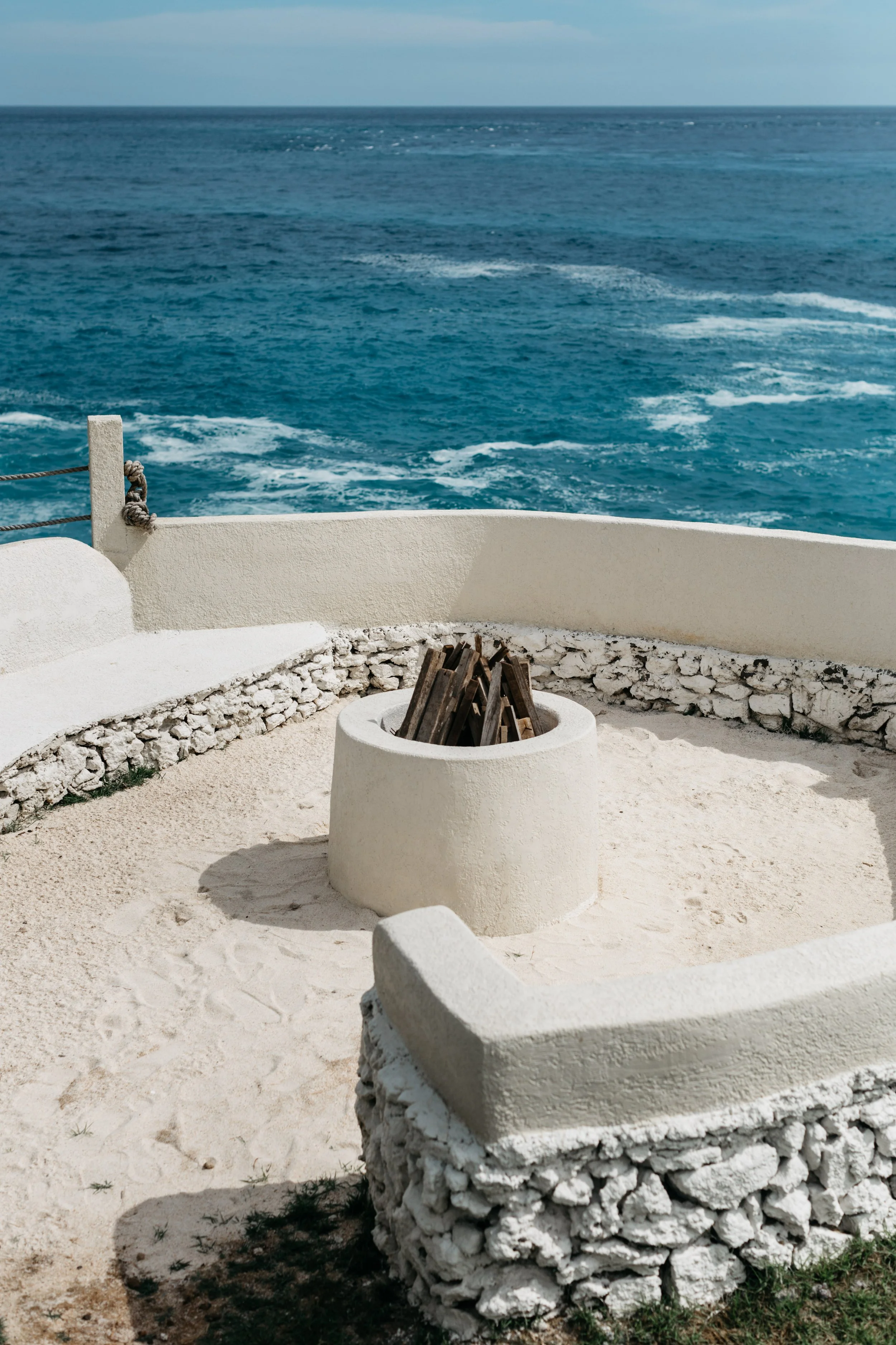 A white stone fire pit on a sandy courtyard overlooks a blue ocean with waves and a cloudy sky.