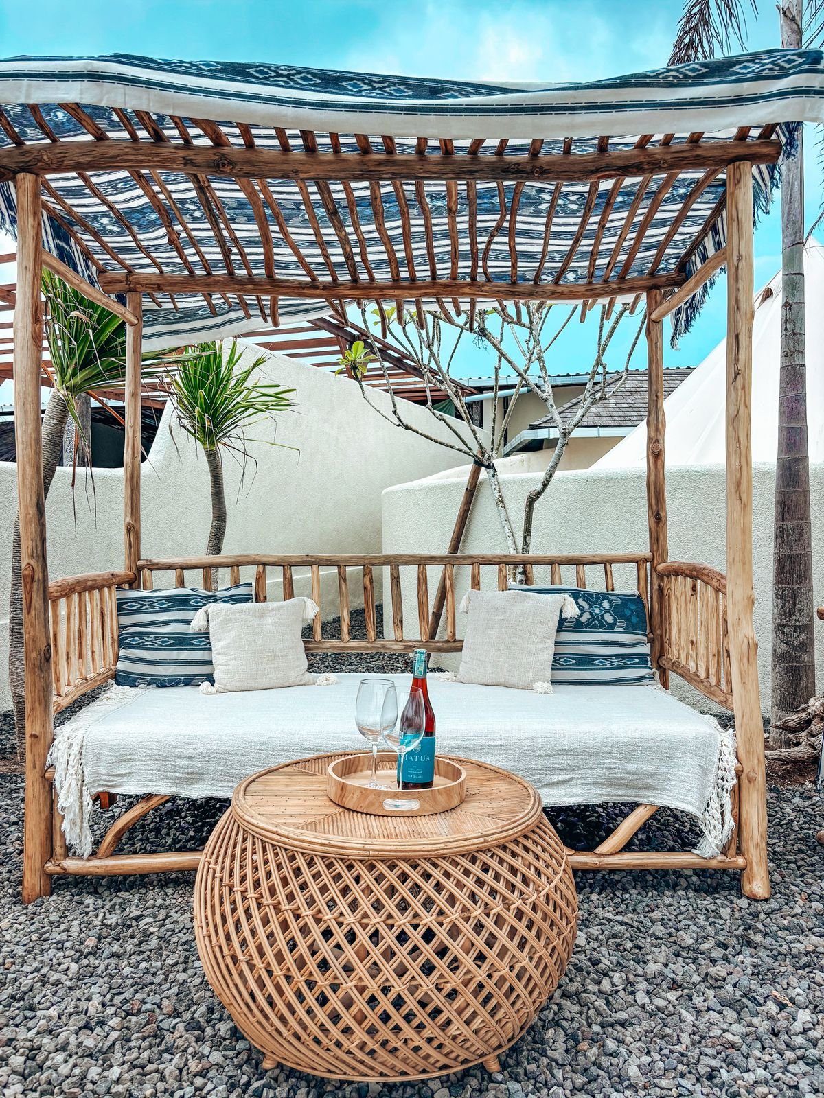 Outdoor seating area with a wooden canopy, white cushion, decorative pillows, a rattan side table with a wine bottle and glass, and tropical plants in the background.