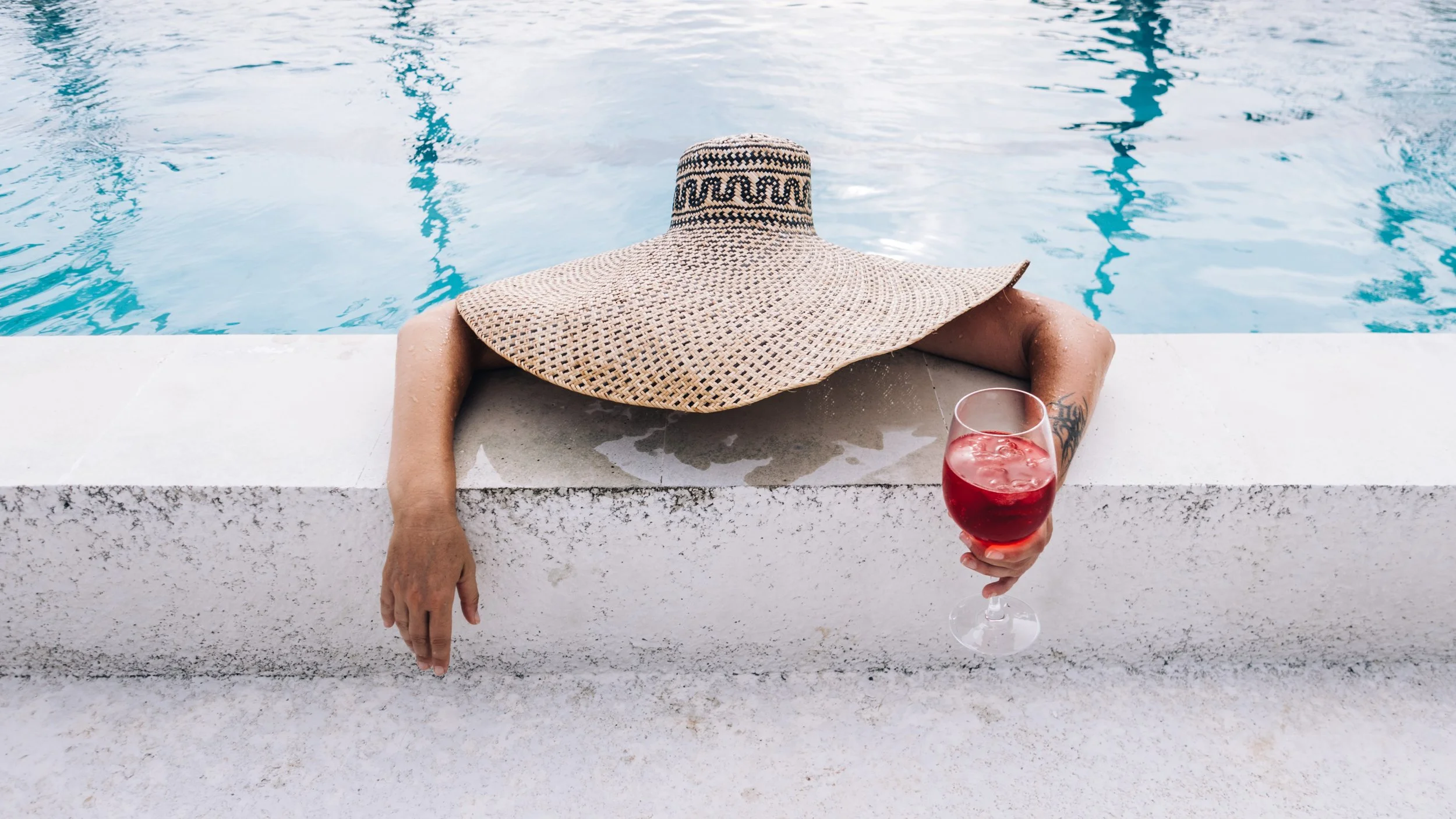 Person relaxing at the edge of a swimming pool, wearing a large straw hat, with one arm resting on the poolside and holding a glass of red wine.