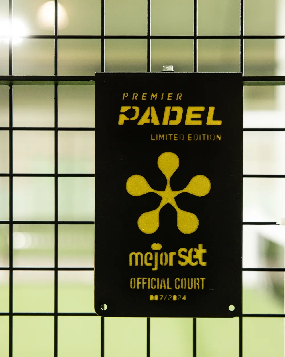 Panneau noir avec écriture jaune indiquant "Premier Padel Limited Edition, meilleur set, Official Court, 07/2024", fixé à une grille en métal.
