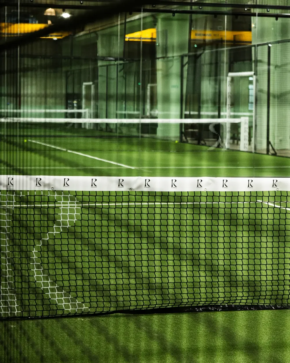 Terrains de padel en intérieur avec un filet au premier plan et des murs en verre derrière.