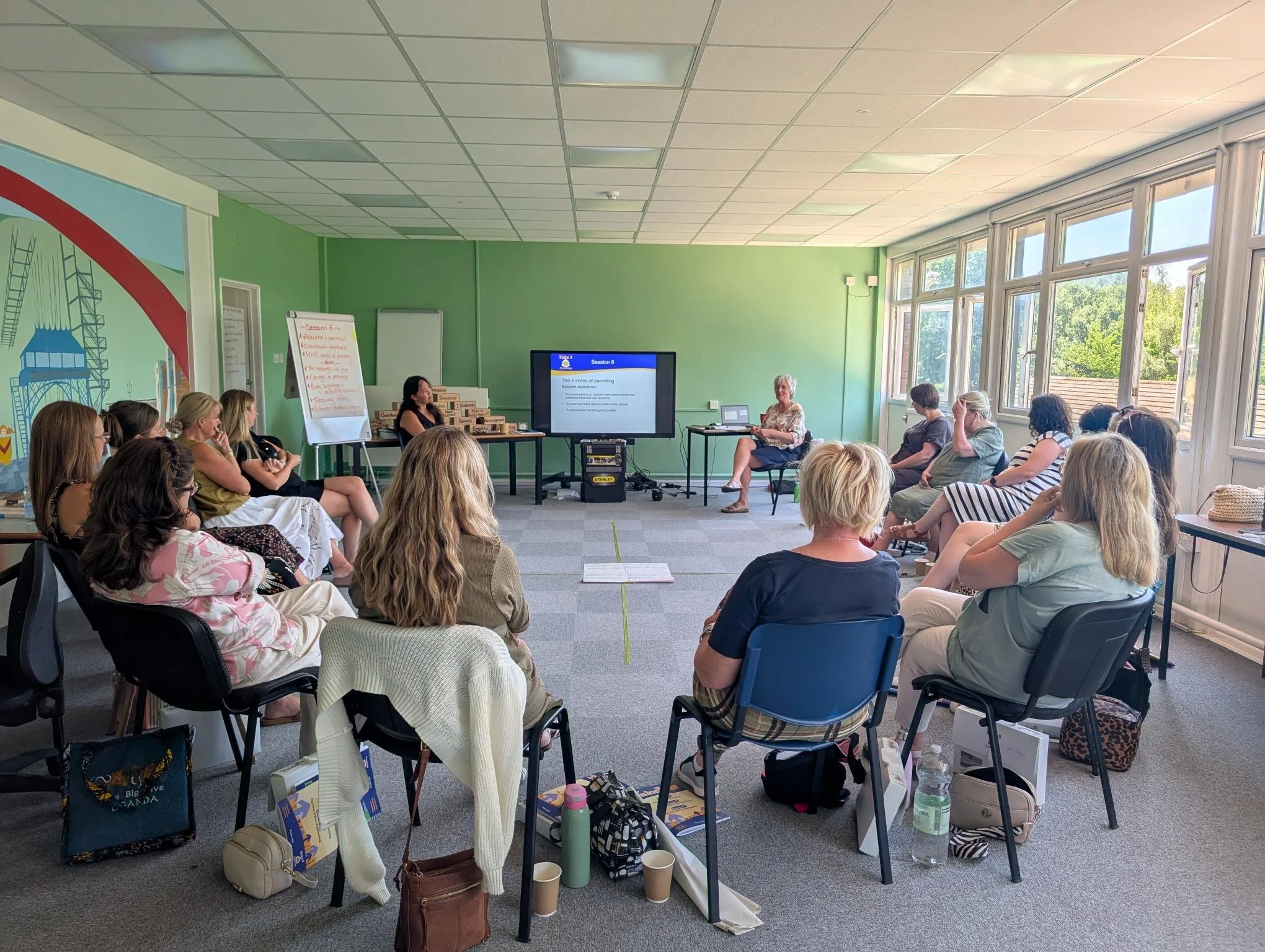 Image shows group of trainees sitting on chairs in a semi-circle layout within a conference room. Two trainers sit at the far end of the room on either side of a projector screen showining text on a powerpoint. There is a flipchart in the far corner.