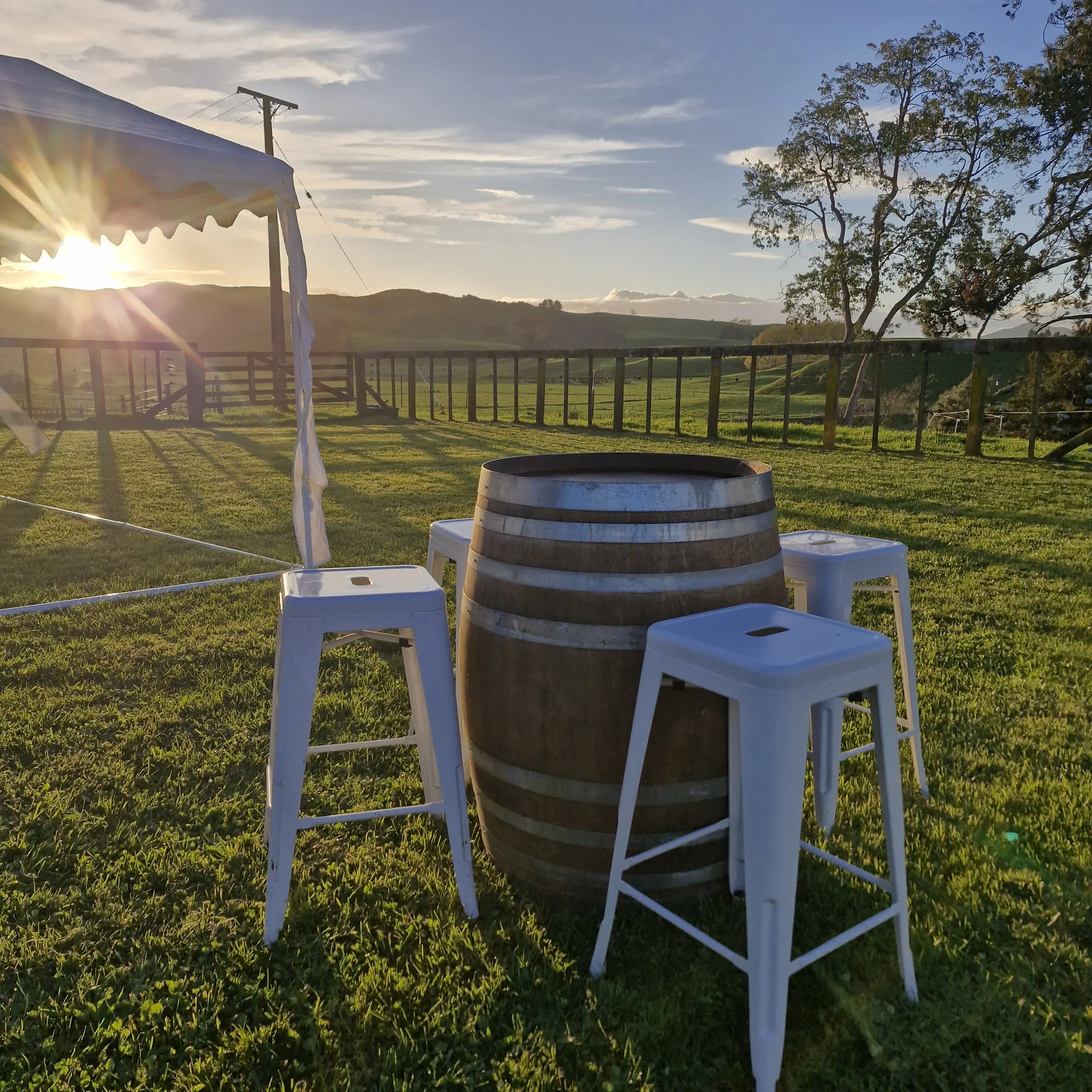 Sunset over a rural landscape with rolling hills, a large tree, a wooden fence, and white plastic chairs in the foreground.